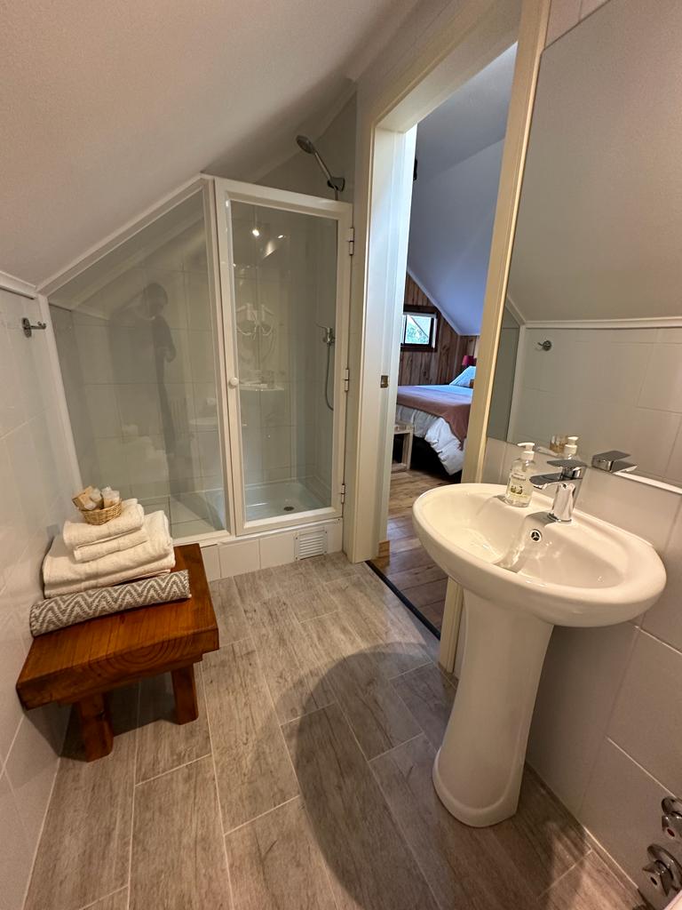 A bathroom is shown featuring a glass shower enclosure and a pedestal sink. A towel set is neatly arranged on a wooden bench, complementing the tiled flooring. Natural light enters the space from the adjacent bedroom, enhancing the peaceful ambiance.