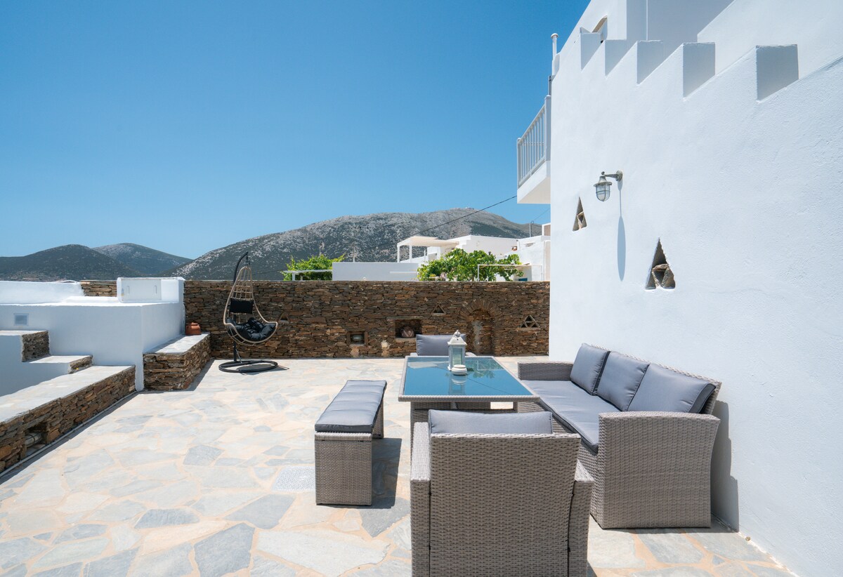 A spacious outdoor patio features a modern seating area with comfortable cushions and a glass table. The surrounding stone wall provides privacy, while the impressive mountain view is visible in the background, complementing the bright blue sky's clear atmosphere.