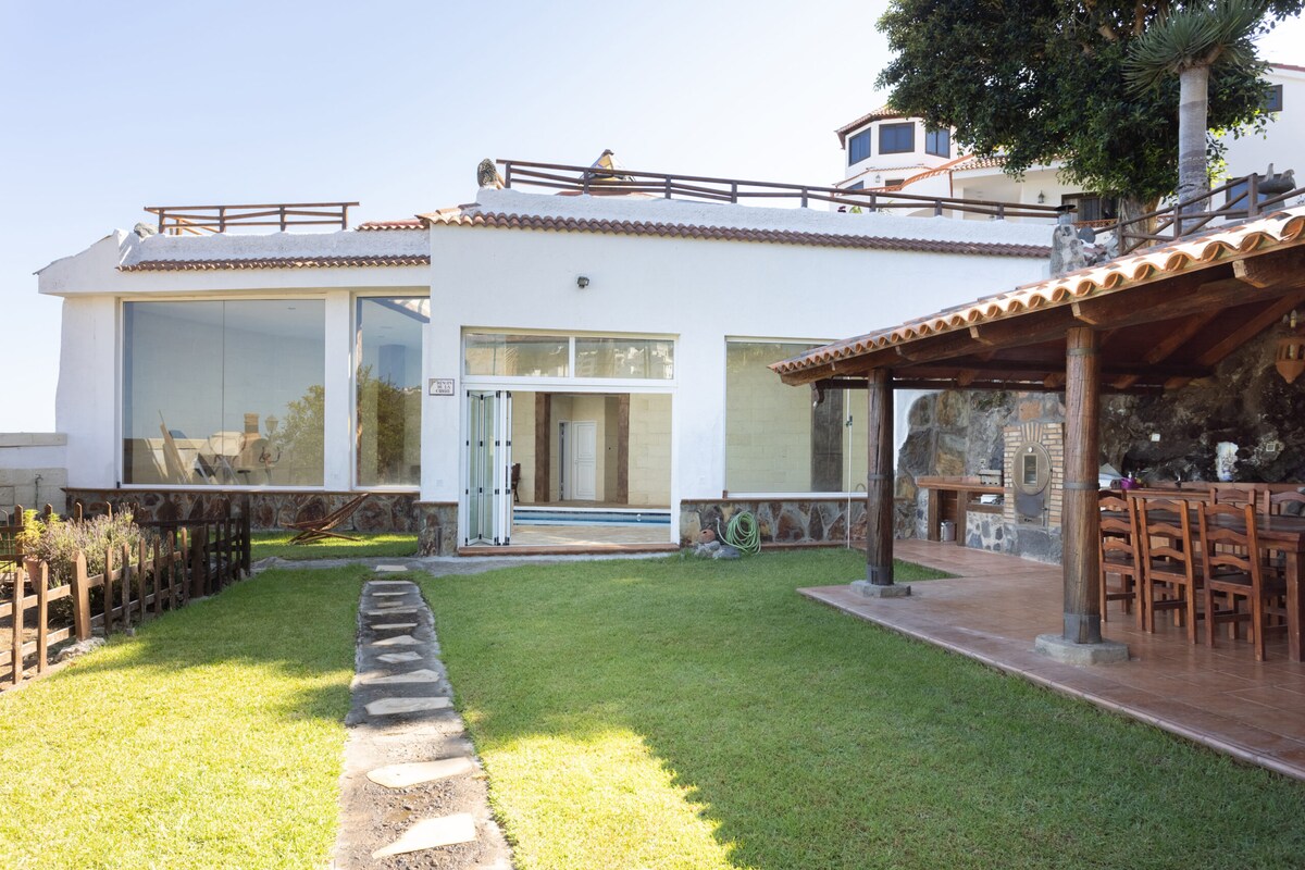 The exterior of the apartment showcases large glass windows overlooking a lush green lawn. The spacious terrace provides an open area for relaxation, while a wooden dining table is placed under a roofed structure, inviting outdoor gatherings. The surrounding garden features neatly arranged stone pathways.