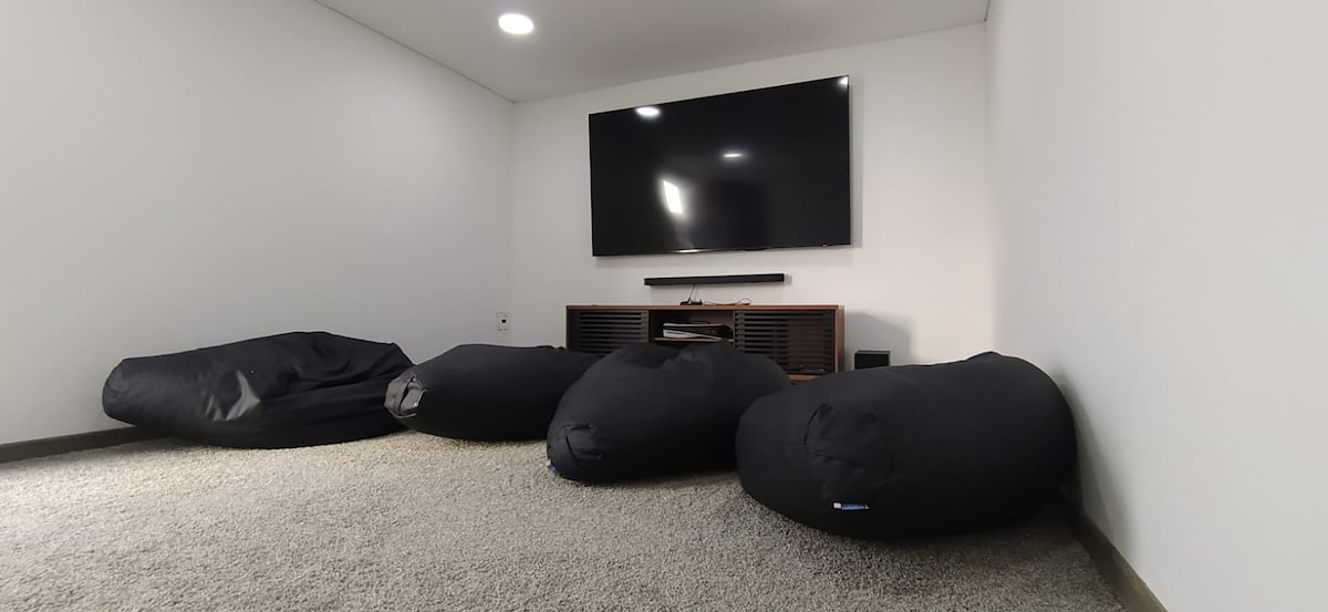 A cozy entertainment area features a large flat-screen television mounted on the wall, accompanied by a low-profile media console. Several black bean bag chairs are arranged on soft carpet, providing a relaxed seating option for guests.