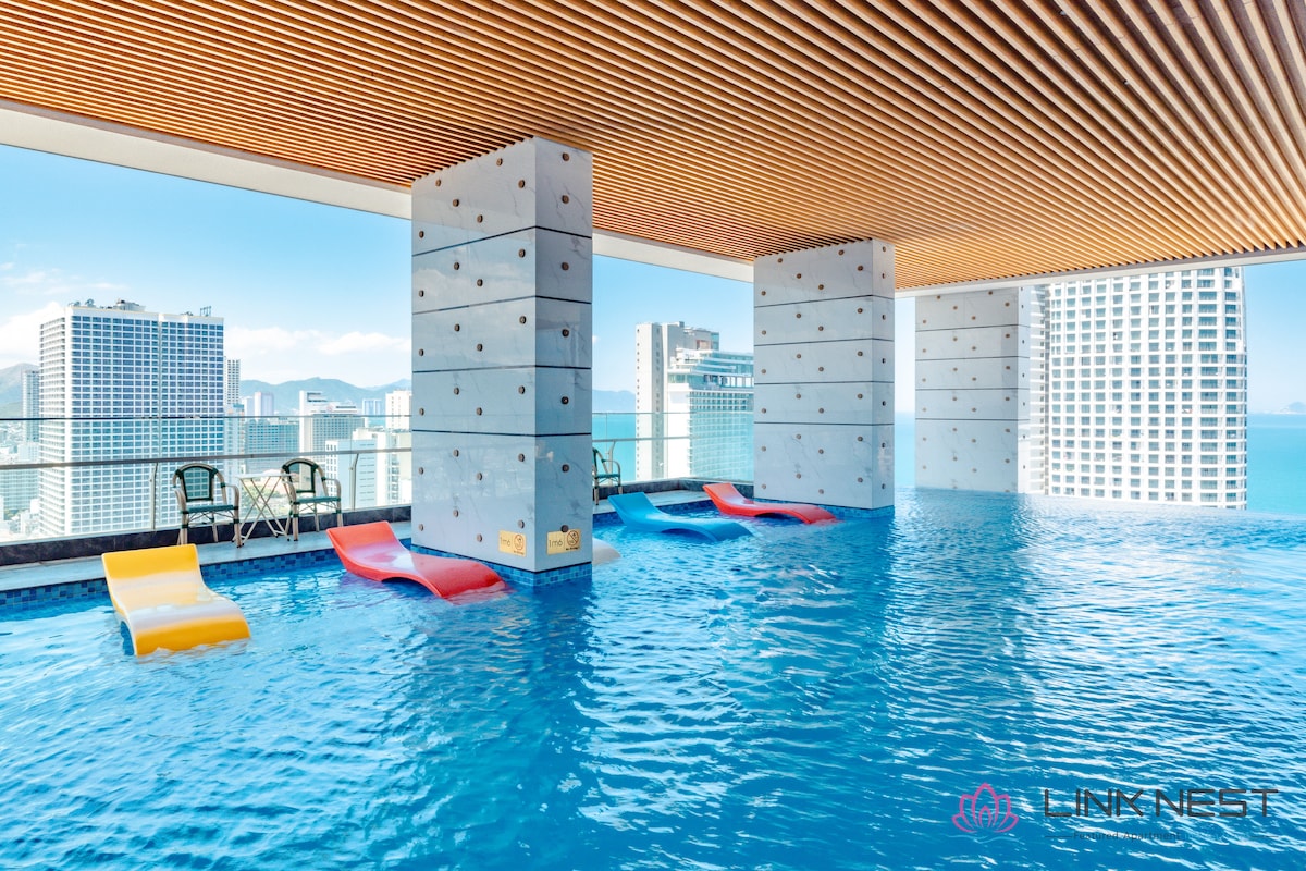 An open-air infinity pool is featured with colorful loungers partially submerged in the water. The surrounding architectural design includes concrete pillars and a wooden ceiling, while breathtaking views of the city skyline and sea can be seen in the background.