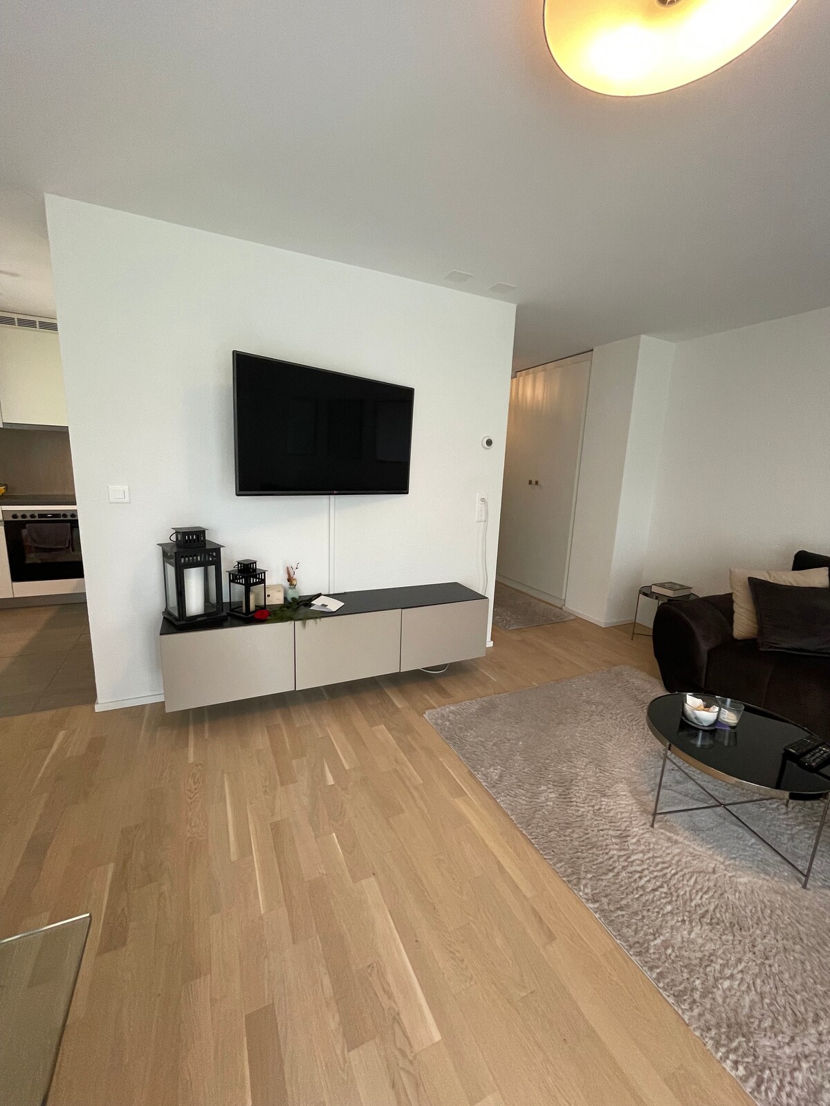 A modern living area features a mounted television above a sleek sideboard. A cozy sofa with cushions is positioned nearby, alongside a round glass coffee table. Natural light fills the space, which has light wood flooring and a soft area rug.