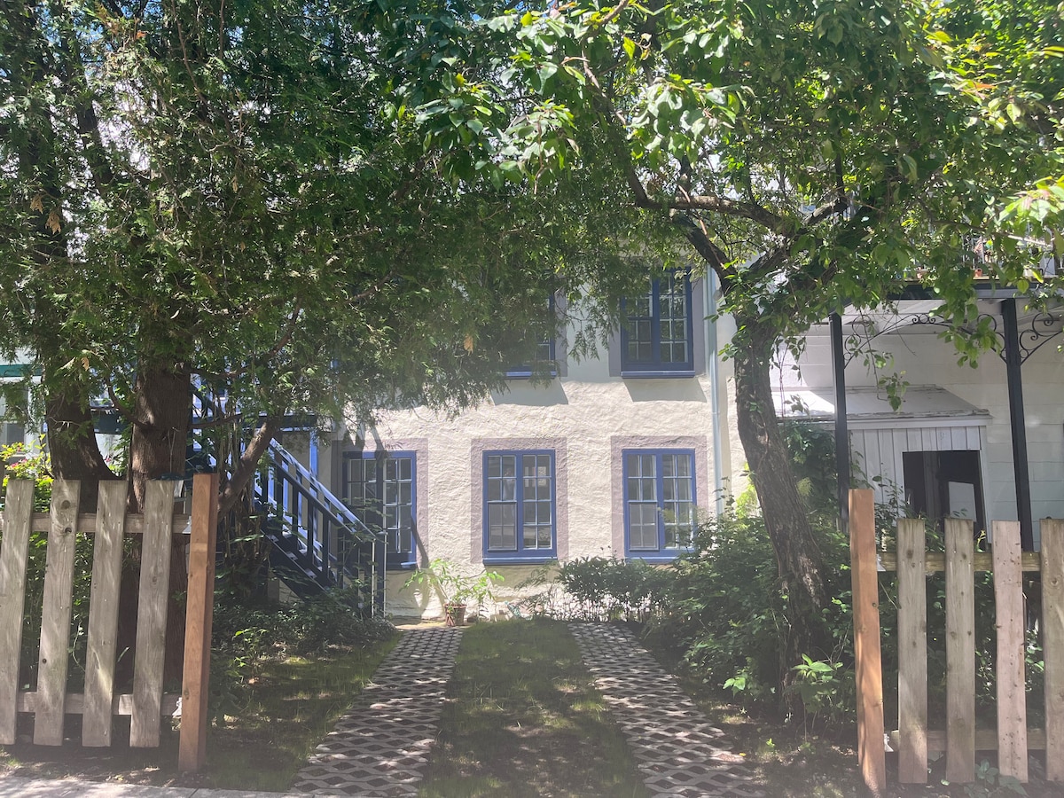 A charming entrance features a stone pathway framed by greenery and wooden fences. The building's exterior showcases light-colored walls with blue window frames. The surrounding trees provide shade, enhancing the tranquil ambiance of the setting.