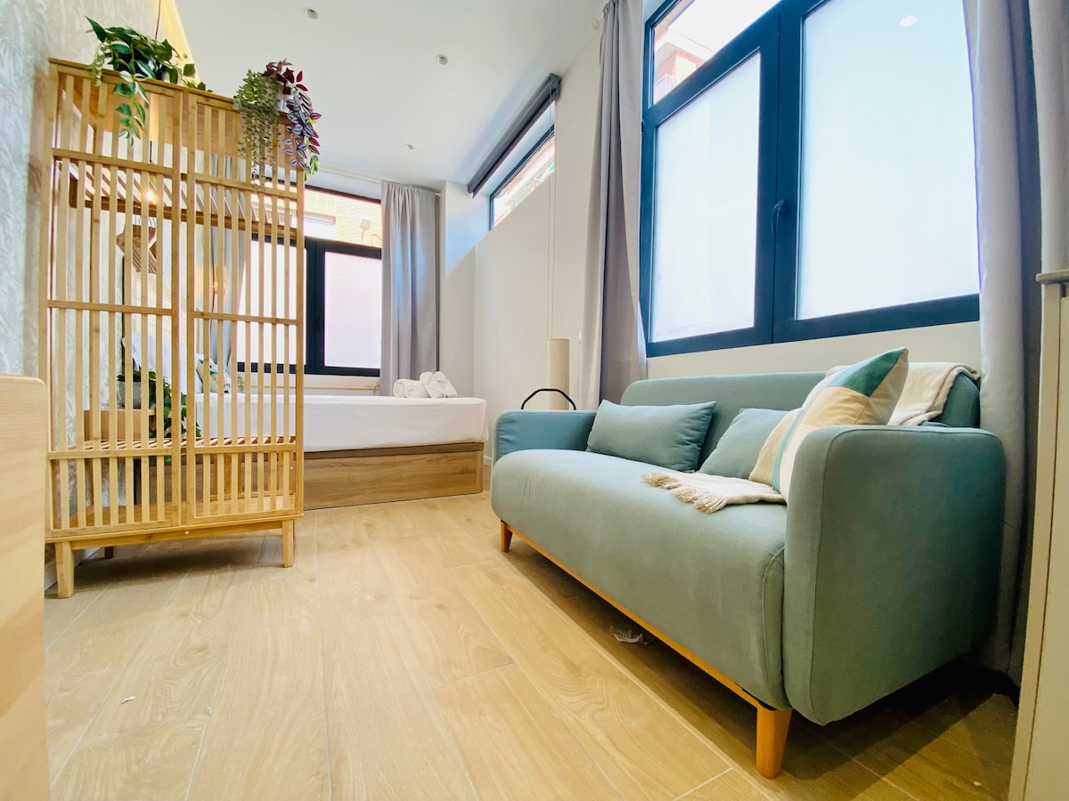 A cozy living space features a light green sofa positioned beside a wooden screen decorated with greenery. Natural light pours through large windows, illuminating the wooden flooring. A spacious bed can be glimpsed in the background, promoting a relaxing atmosphere.