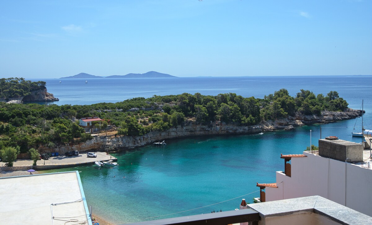 A serene view of the clear blue sea is framed by lush greenery along the shore. The coastline features rocky outcrops and sandy areas, with a few boats gently bobbing in the water. Distant islands can be seen on the horizon under a bright sky.