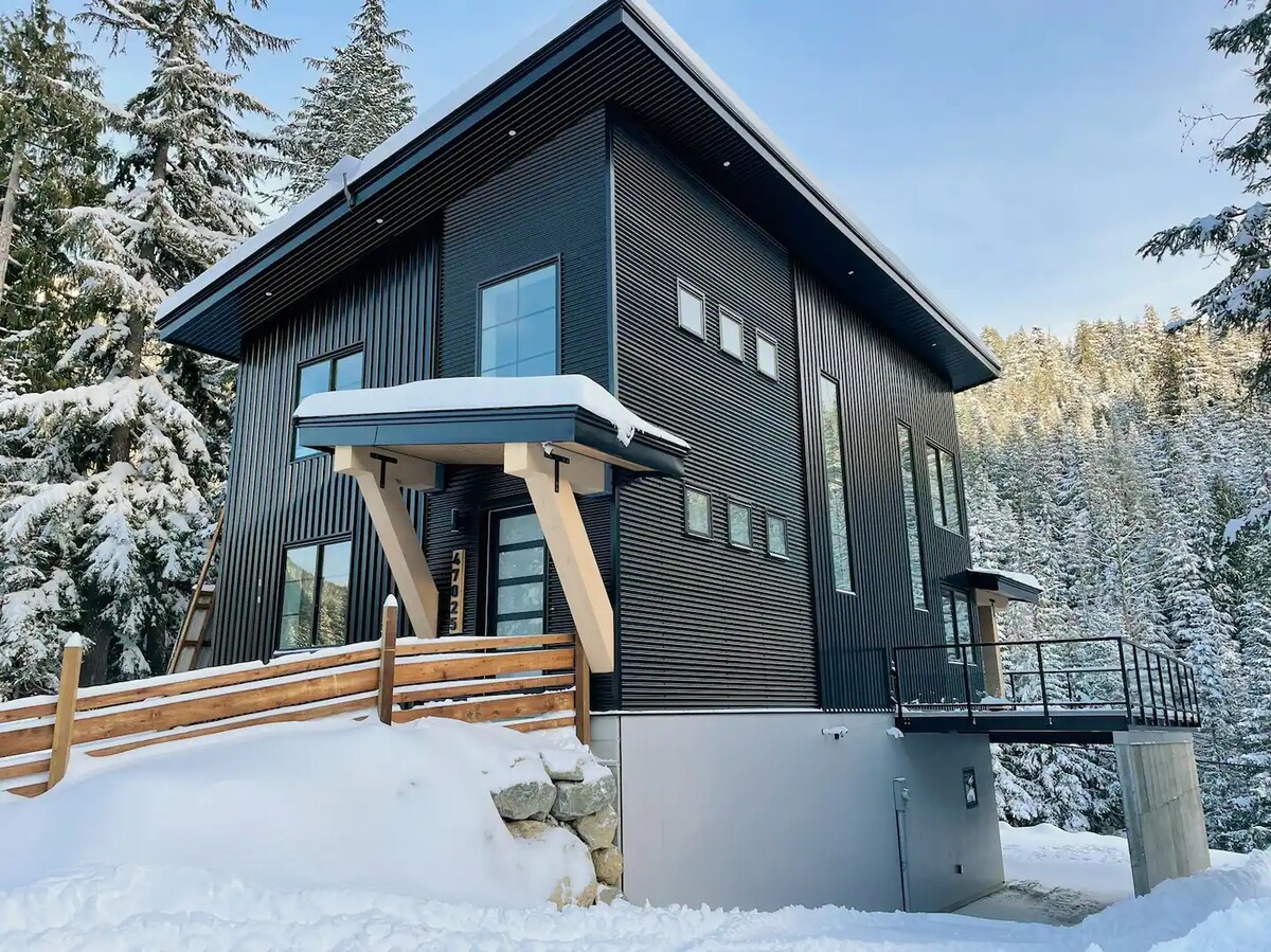 The modern cabin is situated in a snowy landscape, showcasing its sleek black exterior and large windows. An ample deck extends from the building, surrounded by tall trees and a serene winter atmosphere. A snow-covered pathway leads to the entrance.