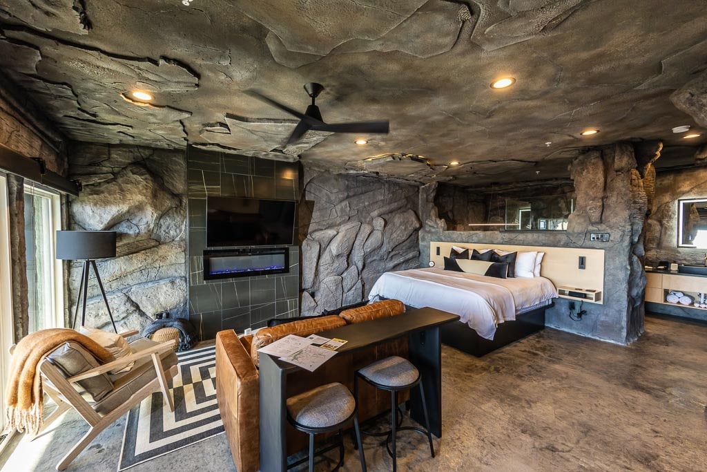 The Cavern Suite features a spacious king-sized bed against a natural rock wall backdrop. A sleek modern fan hangs from the ceiling. A cozy seating area is furnished with a chair and a small table, complemented by a large flat-screen TV mounted nearby.