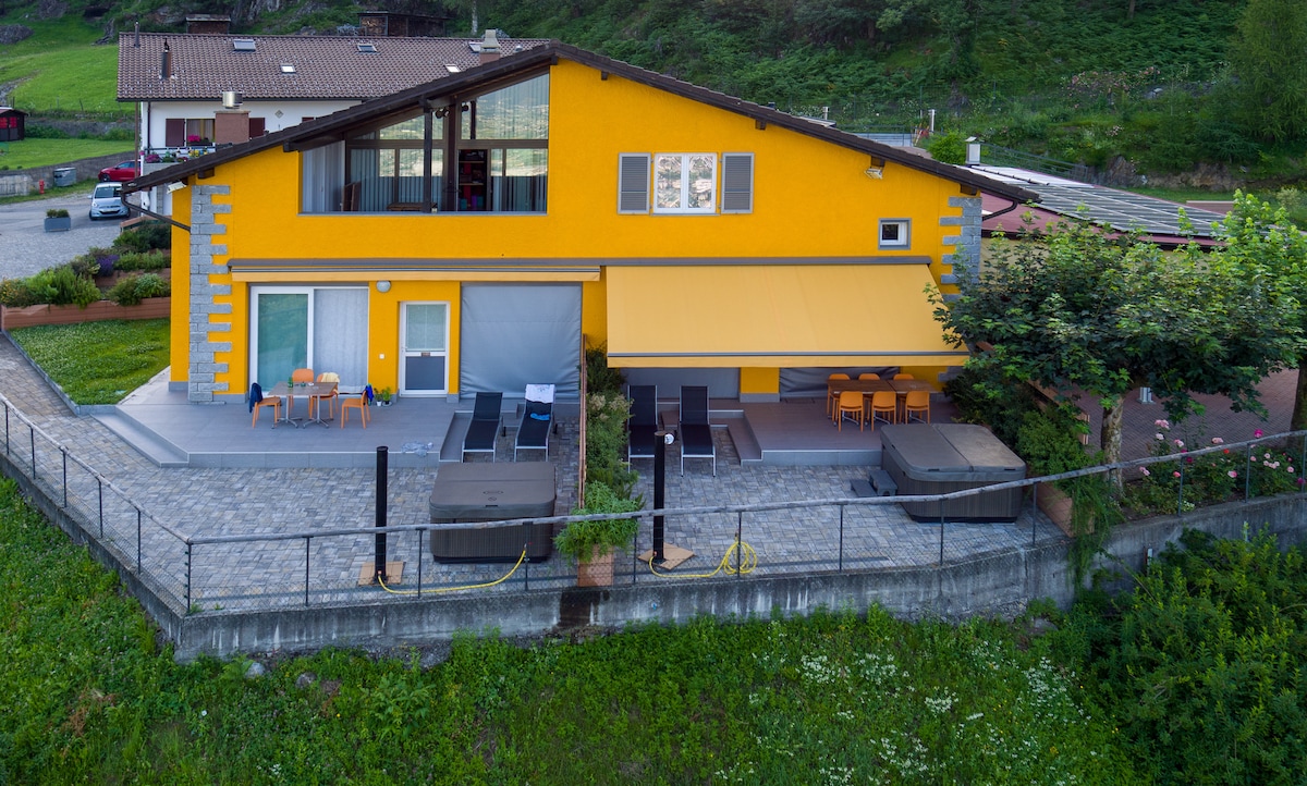 The exterior of a vibrant yellow house is showcased, featuring a spacious terrace equipped with seating areas and lounge chairs. Two hot tubs are positioned near the terrace, surrounded by landscaped greenery, providing a tranquil outdoor space.