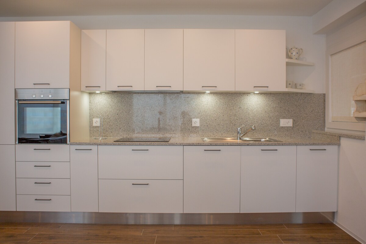 A modern kitchen is featured with sleek white cabinetry and a granite countertop. An oven, stovetop, and sink are integrated into the design. Warm lighting illuminates the workspace, enhancing its functionality, while additional shelves provide storage options above.