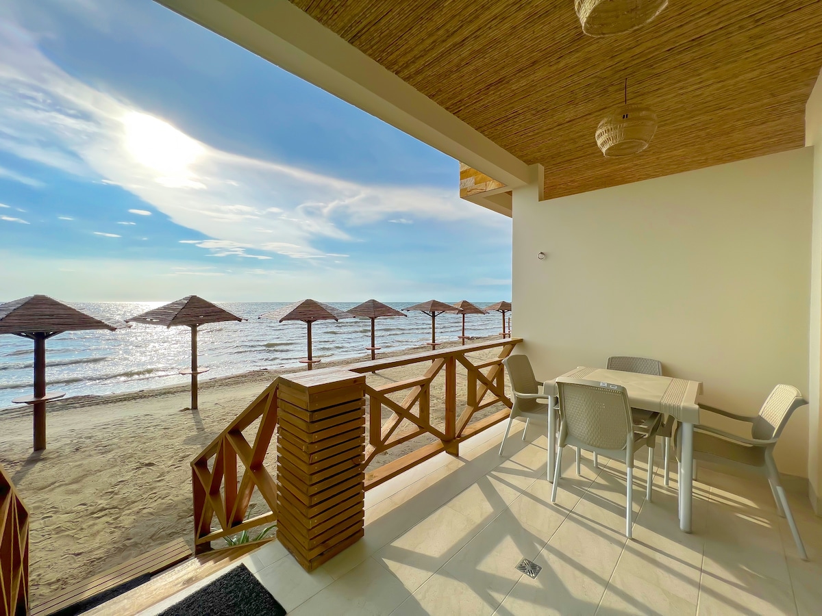 A welcoming outdoor terrace features a dining table surrounded by lightweight chairs, facing the coastline. Sun loungers with umbrellas are positioned on the sandy beach, while the serene sea and sky create a peaceful backdrop.