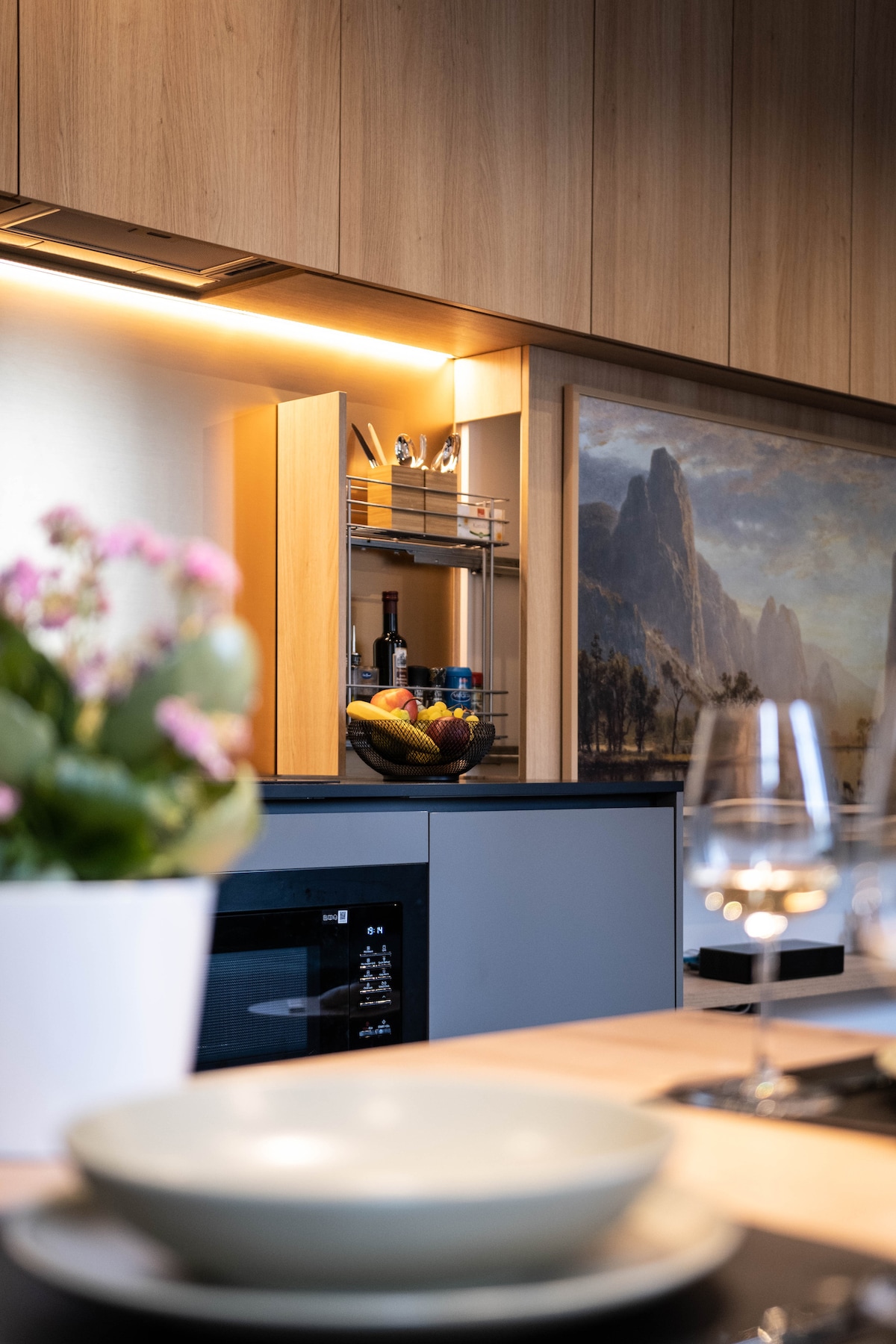 A stylish kitchen area is equipped with modern appliances and storage. A bowl of fresh fruit is placed on the counter, complemented by a potted plant. Subtle lighting enhances the wooden cabinetry, while a scenic painting adds a touch of elegance to the space.