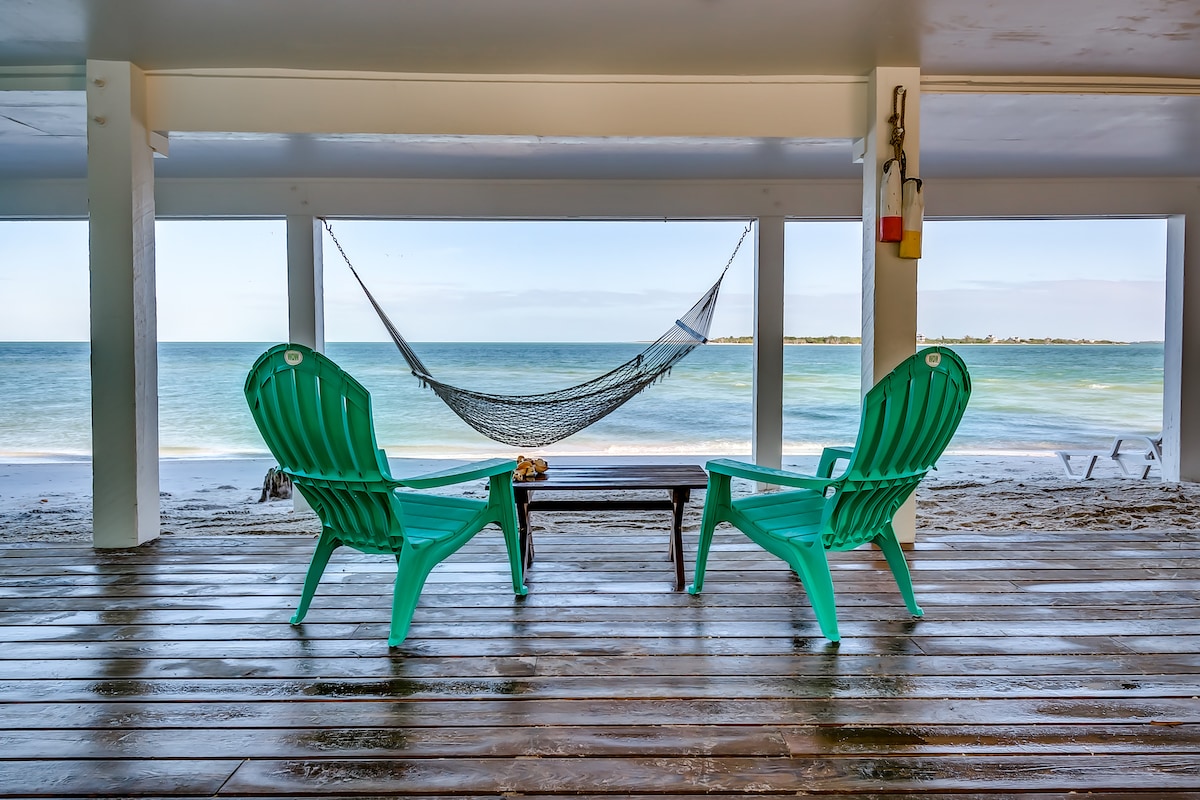 Two green Adirondack chairs are positioned on a wooden deck overlooking the ocean. A small table sits between the chairs, and a hammock is strung nearby. Calm waters and a sandy beach are visible in the distance, enhancing the relaxing coastal ambiance.