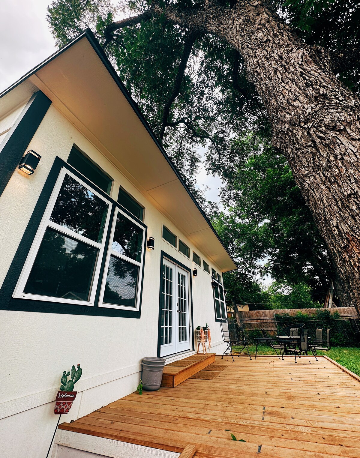 A wooden deck leads to the entrance of the tiny home, framed by large windows. A mature tree provides shade nearby, and a small seating area is visible to the side, inviting outdoor relaxation.
