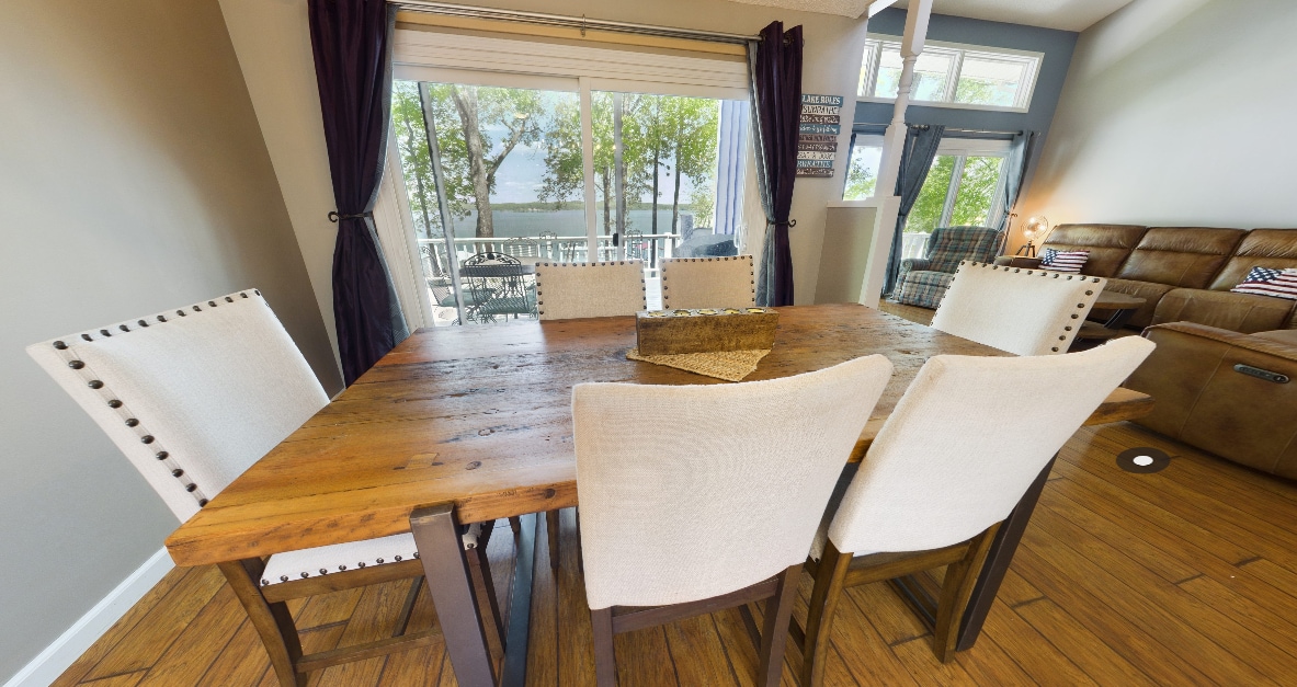 A wooden dining table with a rustic finish is surrounded by six cream-colored chairs. Large windows frame the view of the outdoors, allowing natural light to fill the space. Decorative items are placed at the center of the table, enhancing its inviting appearance.
