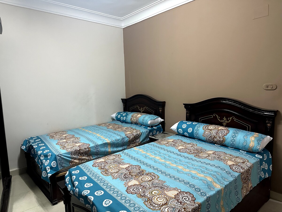 A well-lit sleeping area is equipped with two spacious beds, each adorned with blue and beige patterned bed covers. The dark wooden headboards add a classic touch, and neutral walls create a calm environment.