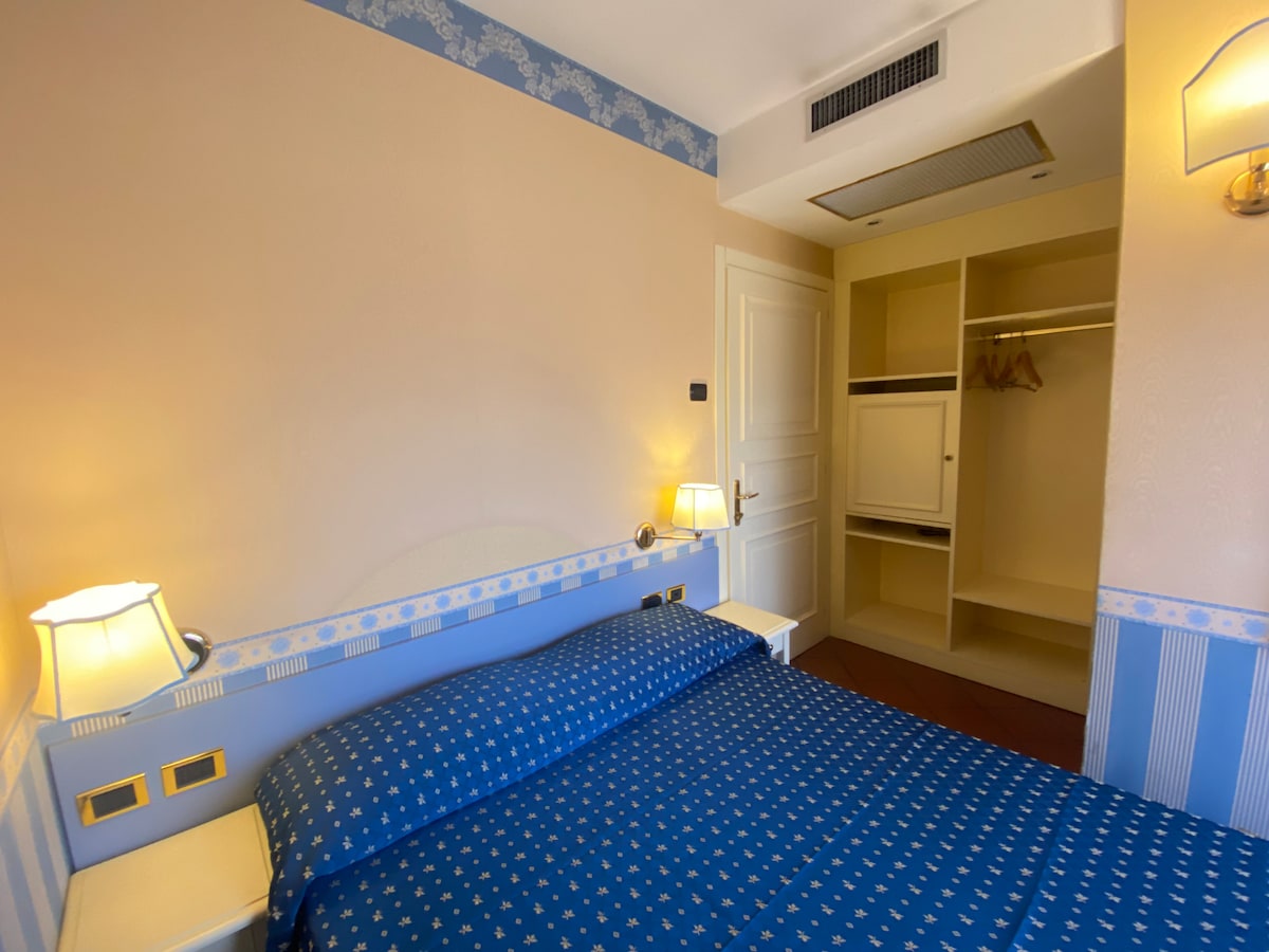 A spacious sleeping area features a blue bedspread patterned with small motifs. The soft light from two bedside lamps illuminates the room. A closet with hanging space and shelves is visible, alongside a minimalistic white built-in unit.