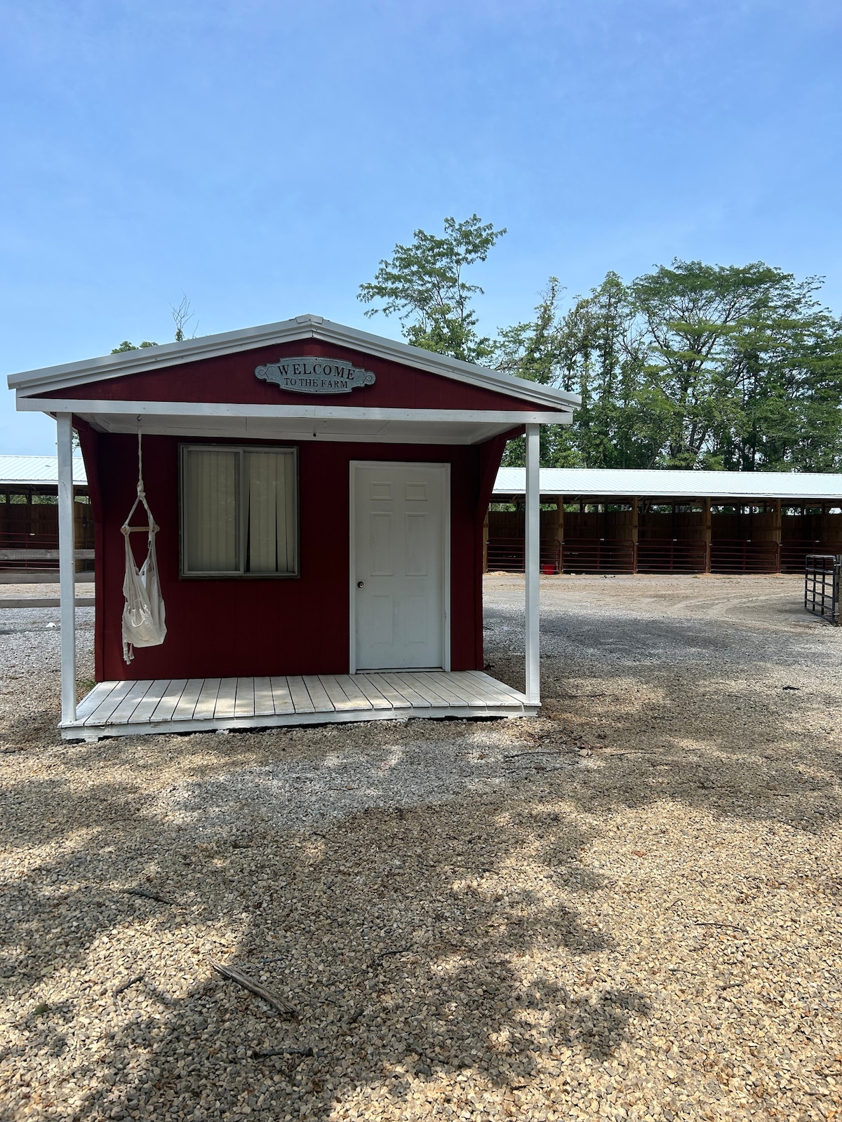A charming red cabin is shown with a white door and a welcoming sign above. A small white swing hangs from the porch, which is surrounded by gravel pathways and trees in the background. Stalls or buildings are visible in the far distance.