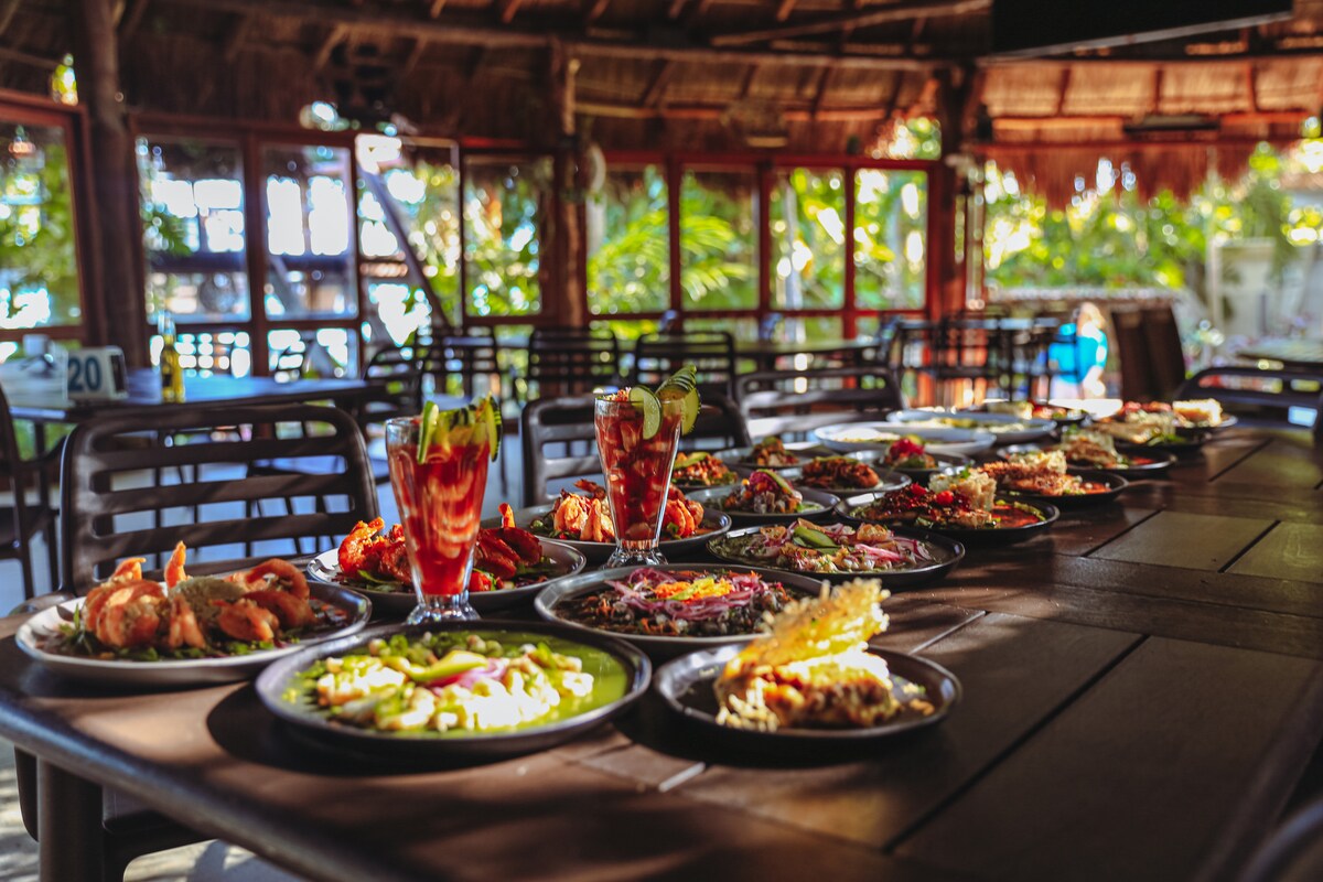 A dining area is filled with a variety of colorful dishes served on black plates. Two refreshing cocktails are displayed prominently among the food, with vibrant garnishes. The open space allows natural light to illuminate the setting, complemented by a view of greenery outside through large windows.