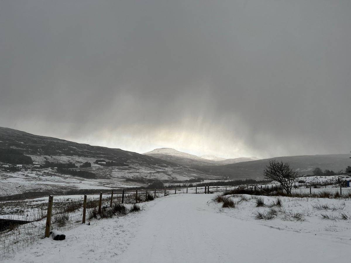 A snow-covered pathway stretches into the distance, surrounded by rolling hills. Soft light filters through dark clouds, illuminating the mountain landscape in the background. Sparse vegetation and a fence line are visible along the path, enhancing the tranquil winter scene.