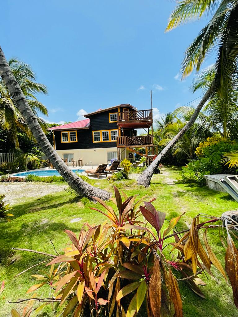 A charming retreat is framed by swaying palm trees, with a lush garden featuring colorful foliage. A private pool is partially visible, complemented by sun loungers for relaxation. The two-story building showcases a vibrant exterior, highlighted by large windows and a balcony overlooking the grounds.
