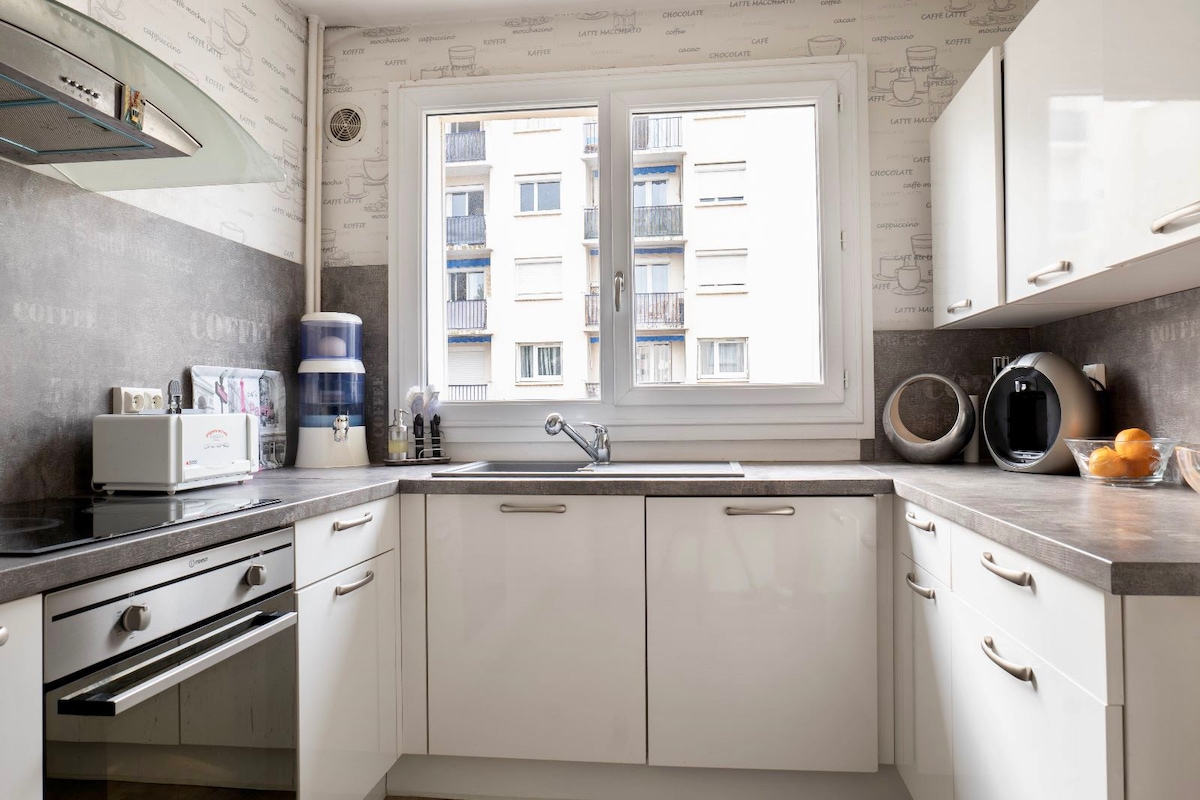 A modern kitchen is equipped with white cabinetry and a gray countertop. A window allows natural light to fill the space while offering a view of the exterior. Appliances include an oven, a microwave, and a kettle. A water dispenser and fruit are visible on the countertop.