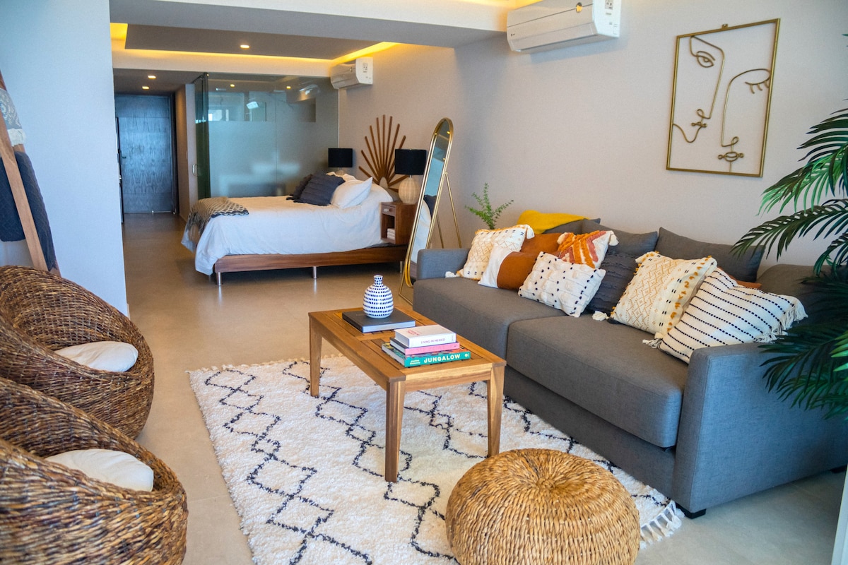 A spacious living area features a comfortable grey sofa adorned with a variety of decorative cushions. A wooden coffee table sits in the center, accompanied by a woven pouf. Two elegant armchairs are placed near the wall, complementing the decor with a sense of warmth.