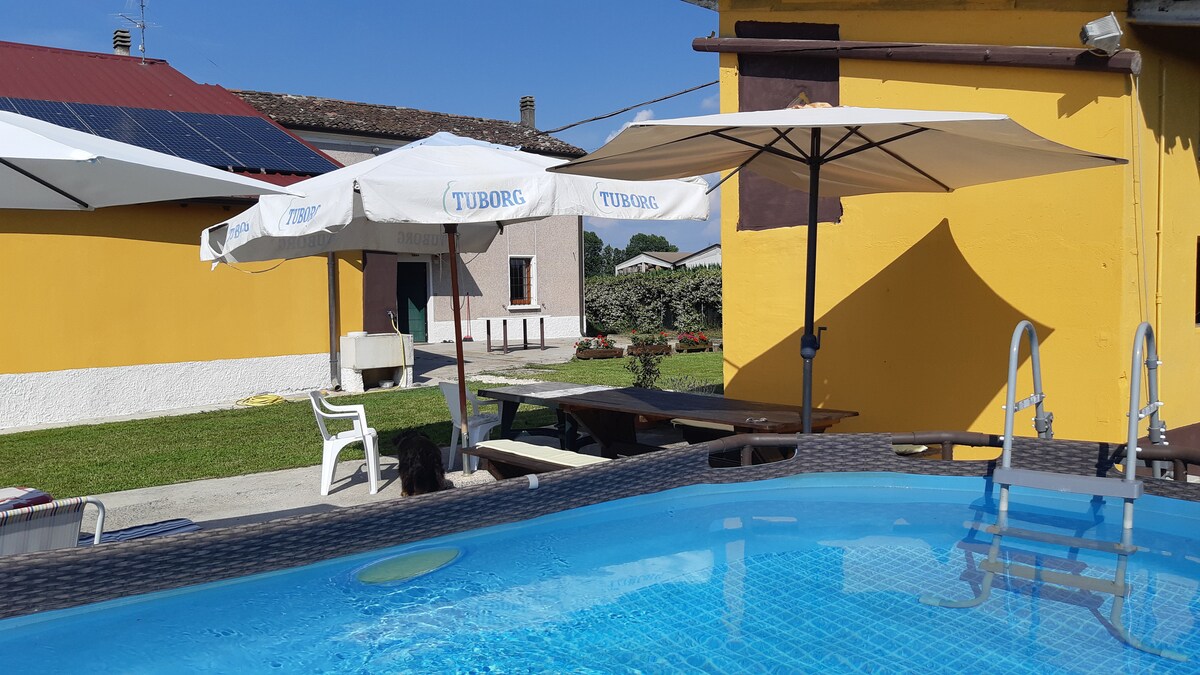 A private outdoor space is highlighted by a clear, above-ground pool with a ladder, surrounded by sun loungers and umbrellas. The vibrant yellow exterior of the house creates a cheerful backdrop, while picnic tables and green lawn area are visible in the distance.