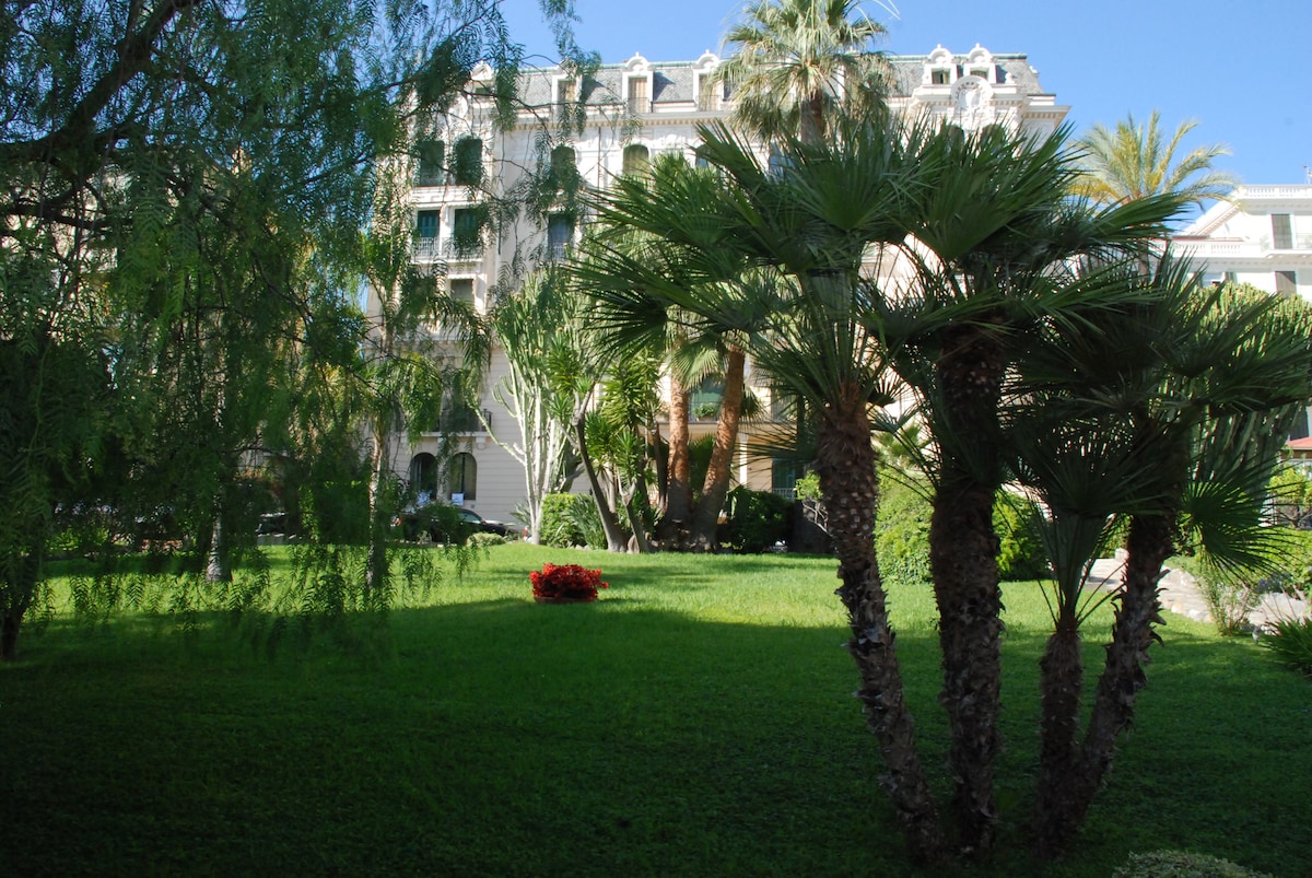 A lush garden area is depicted, featuring well-maintained green grass and several palm trees. Bright flower beds add color to the landscape, while classic villa architecture can be seen in the background, contributing to the peaceful outdoor environment.