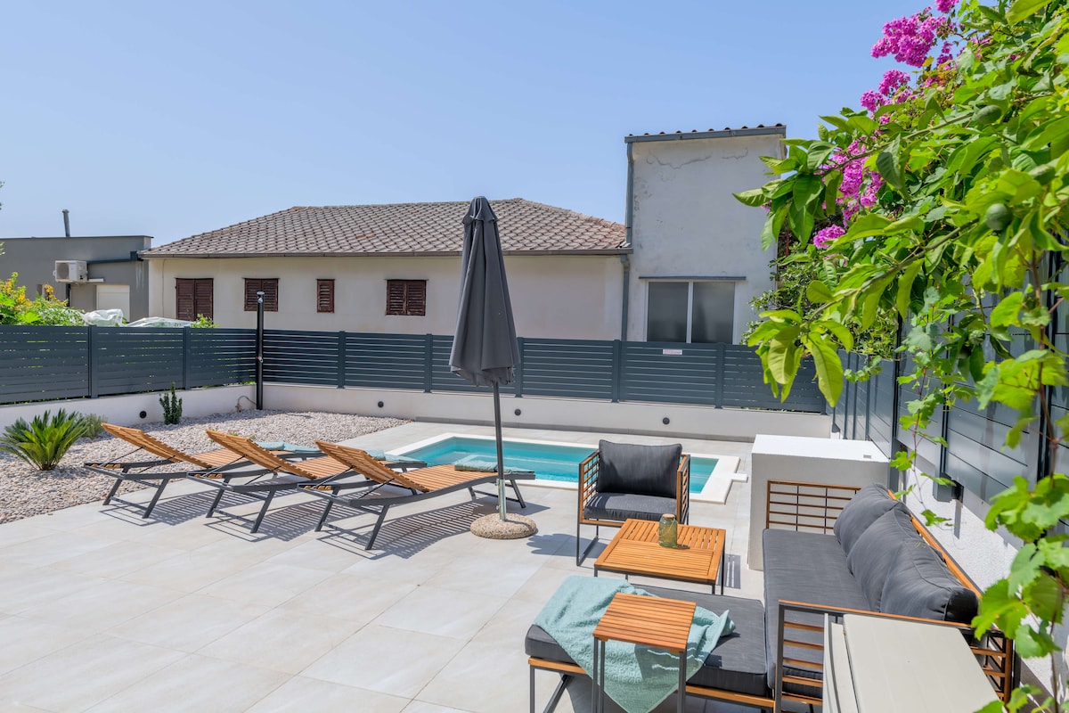 An outdoor area features a refreshing rectangular pool surrounded by a spacious paved terrace. Several lounge chairs are positioned for relaxation, alongside a seating area with modern furniture. Lush greenery and flowering plants add a touch of color to the setting, with a large umbrella providing shade.
