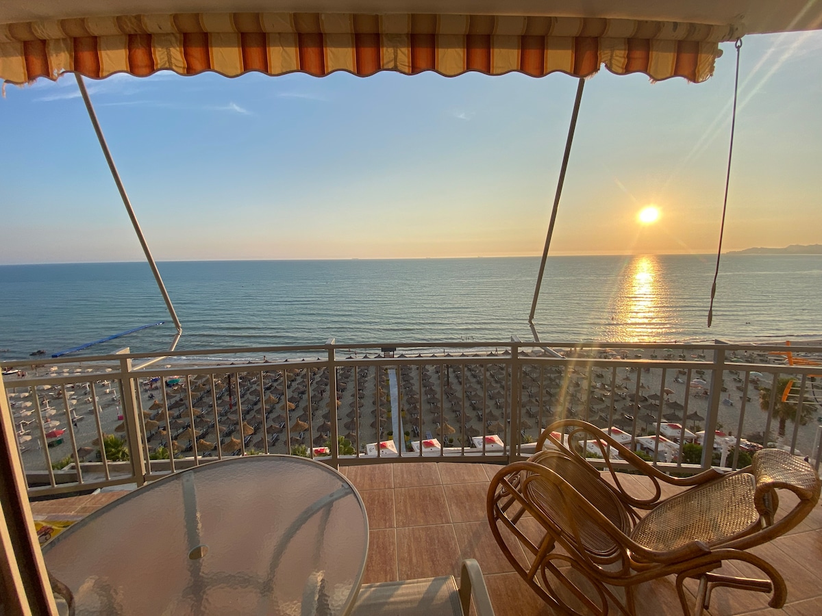 A spacious balcony offers stunning views of the ocean and sunset. The scene features a round glass table and wicker chairs. Below, a beach with numerous sun umbrellas and lounge chairs can be seen, providing a sense of relaxation and seaside enjoyment.