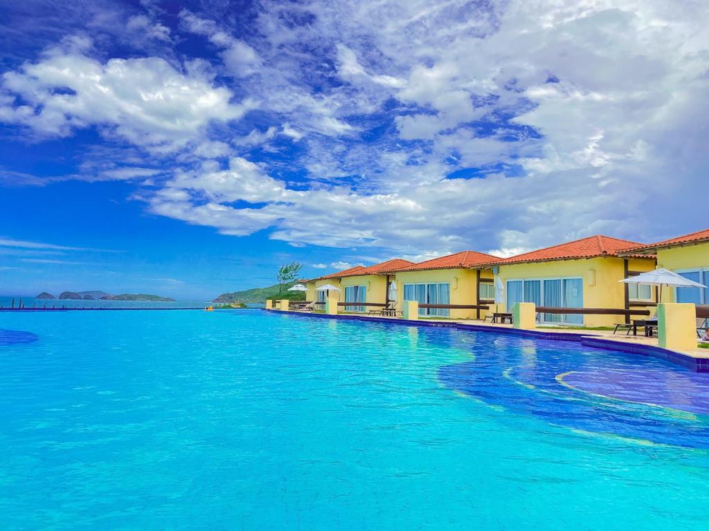 A vibrant pool area is showcased, seamlessly merging with the horizon. The calm surface reflects the blue sky and scattered clouds. Spacious bungalow accommodations can be seen along the pool's edge, featuring large windows and lounging areas, enhancing the relaxing atmosphere.