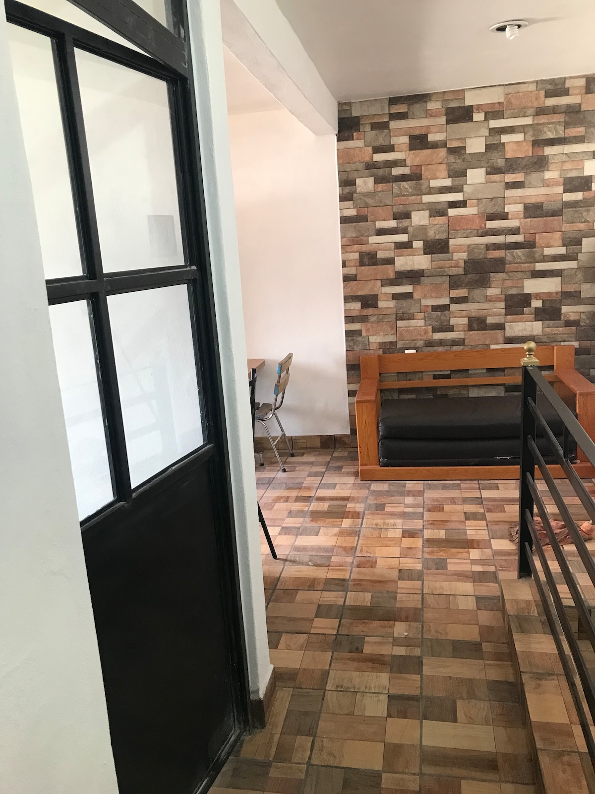 A view from a doorway reveals a cozy seating area with a wooden frame sofa, complemented by a textured accent wall featuring various earthy tones. Smooth tile flooring extends throughout the space, leading towards a small dining area with metal chairs visible in the background.