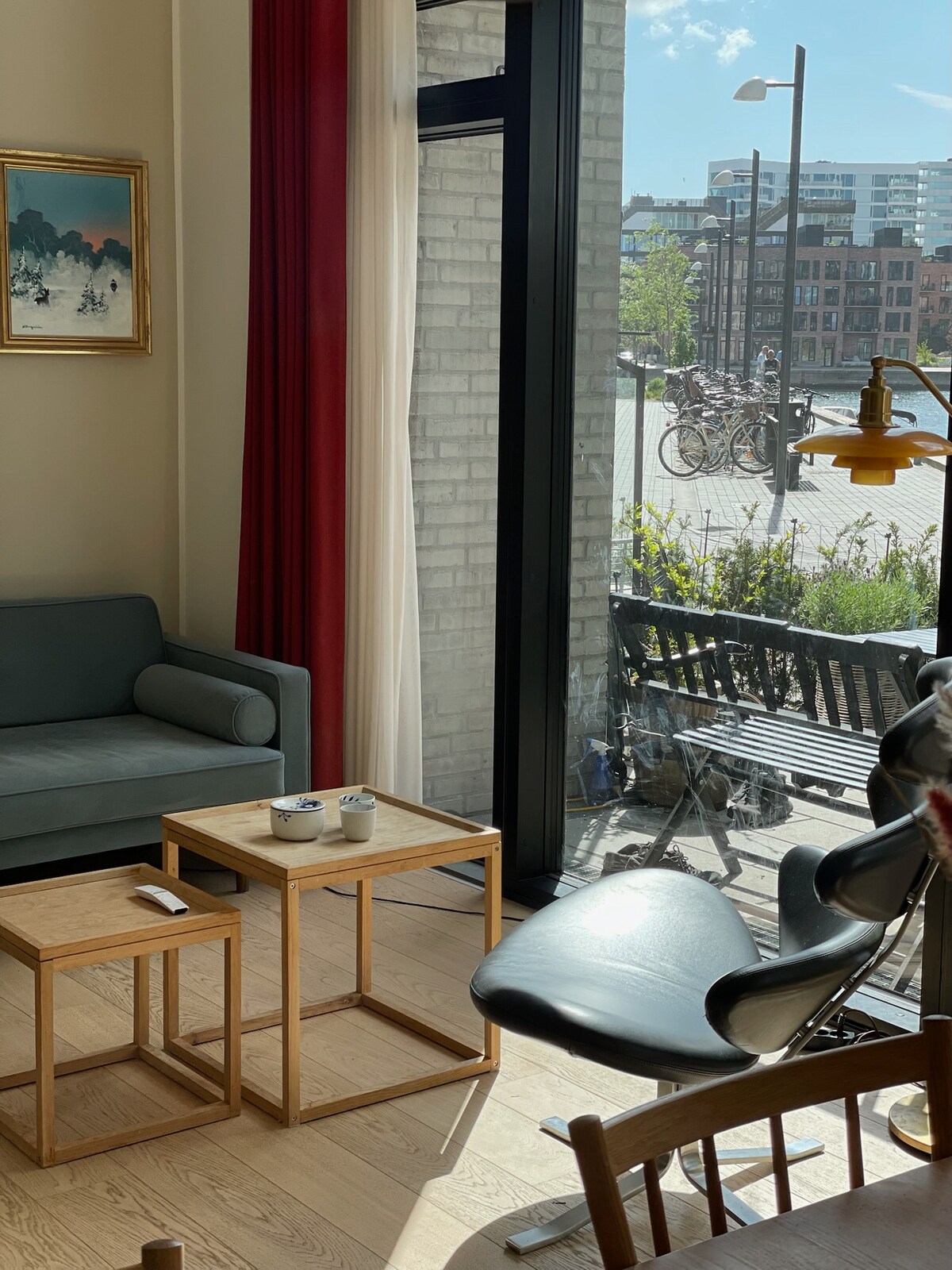 A bright living area is seen, featuring a gray sofa and two wooden side tables. Natural light filters through the large glass doors, illuminating the space. Outside, a view of bicycles and greenery is visible, enhancing the connection with the outdoors.