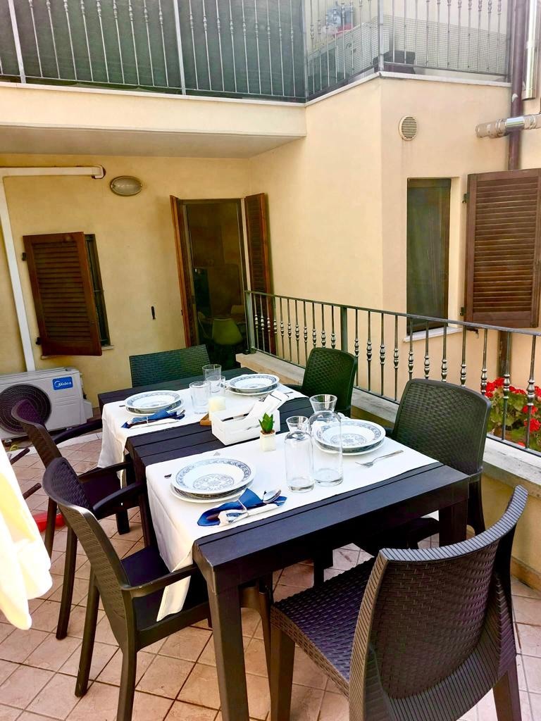 An outdoor dining area features a dark table set for six, with plates and glasses arranged neatly. The space is enhanced by surrounding greenery and offers a welcoming atmosphere for meals. Windows with wooden shutters and a railing are visible in the background.