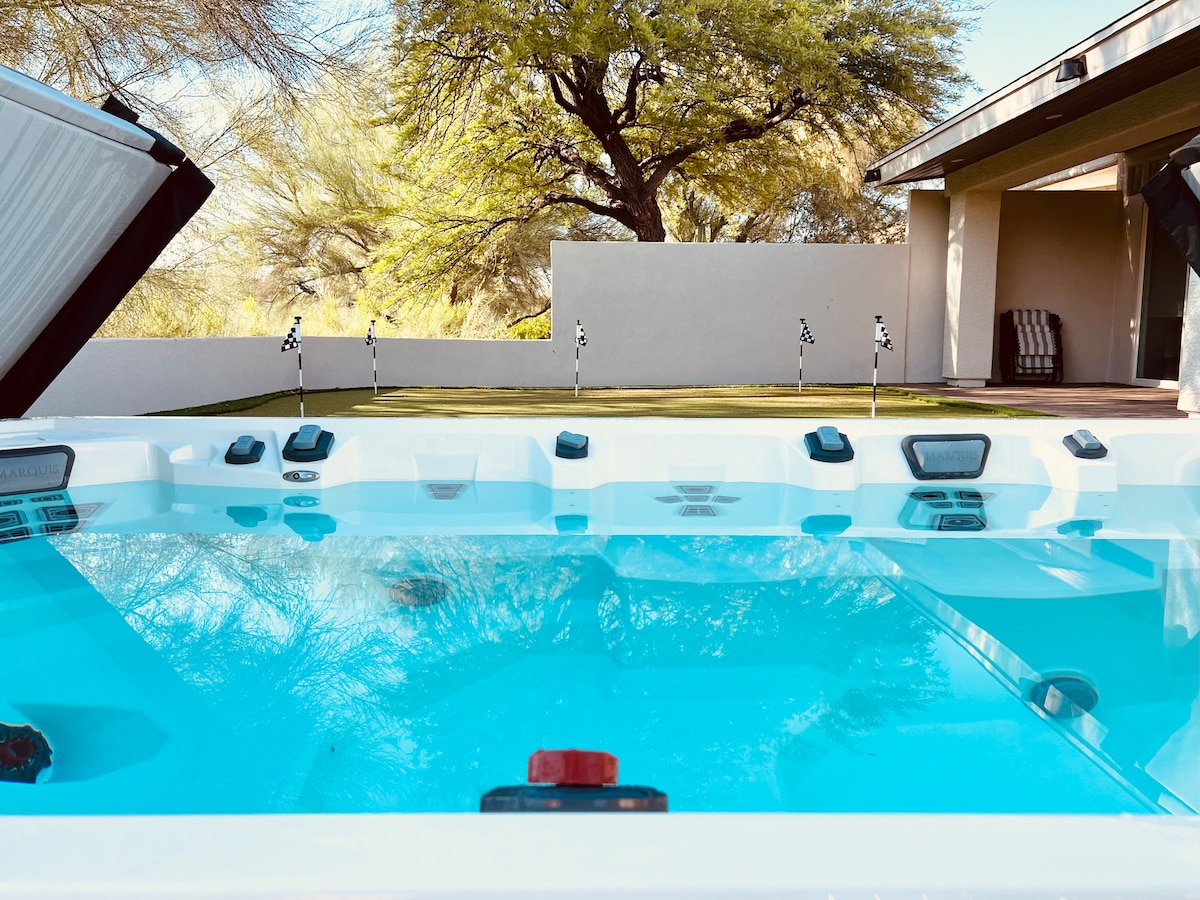 A large swim spa is visible, filled with clear blue water. Massage seats and control settings are integrated for aquatic fitness. In the background, a spacious green area is seen, lined with putting green flags and shaded by trees.