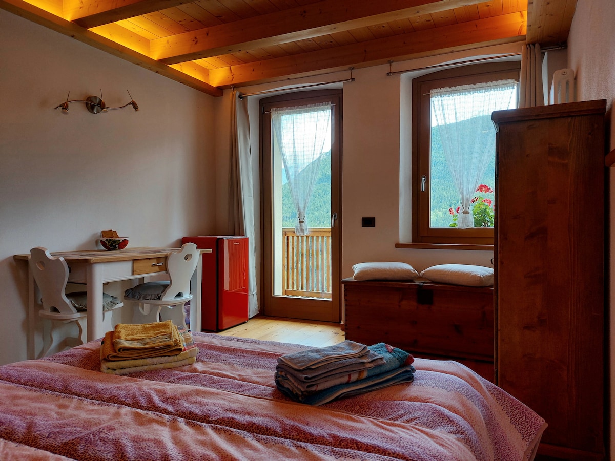 A well-lit room features a spacious bed with neatly arranged towels. A small white table is positioned next to a red refrigerator. Large windows reveal a view of the mountains, framed by light curtains. A wooden trunk adds character to the cozy atmosphere.