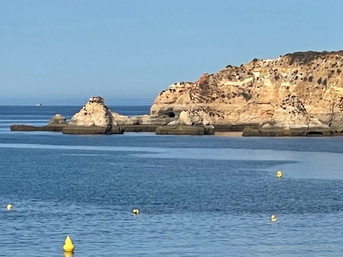 A rocky coastline is visible against a calm sea under clear blue skies. The formations are outlined by the gentle waves, with yellow buoys floating in the foreground, marking the water. The scene conveys a serene coastal environment.