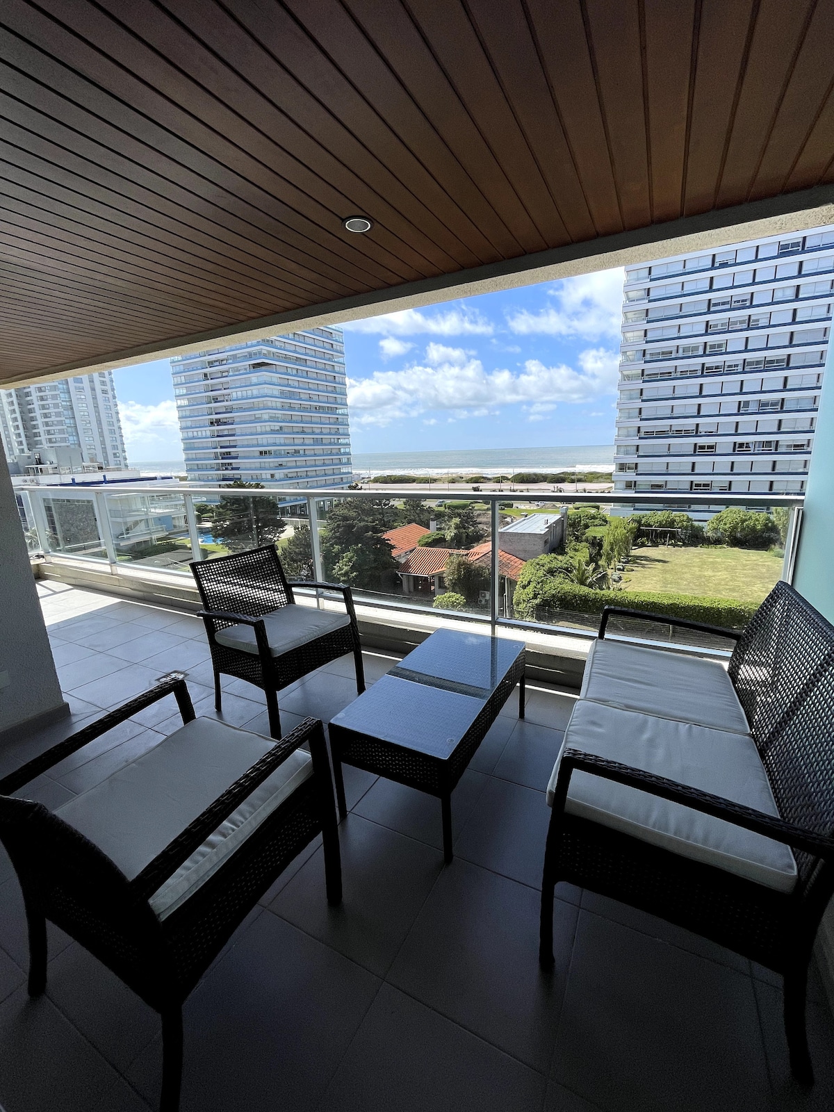A furnished balcony space is visible, featuring two chairs and a coffee table. A view of the greenery and nearby buildings is framed by a glass railing, with the ocean visible in the distance under a partly cloudy sky.