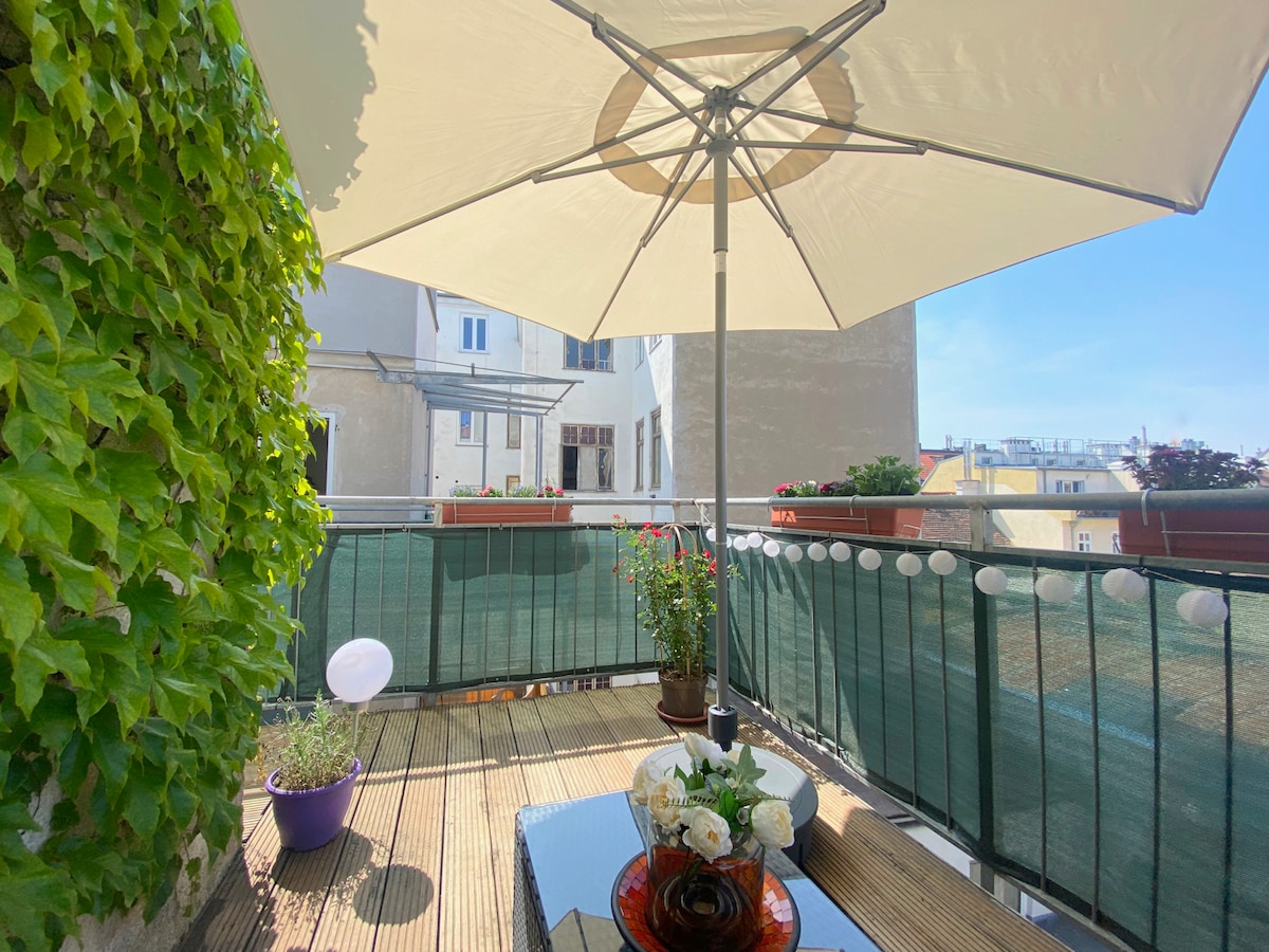 A sunny balcony is shaded by a large umbrella, featuring a small table adorned with a decorative plate and flowers. Lush greenery borders the space, and potted plants add color, while the view showcases adjacent buildings under a clear blue sky.