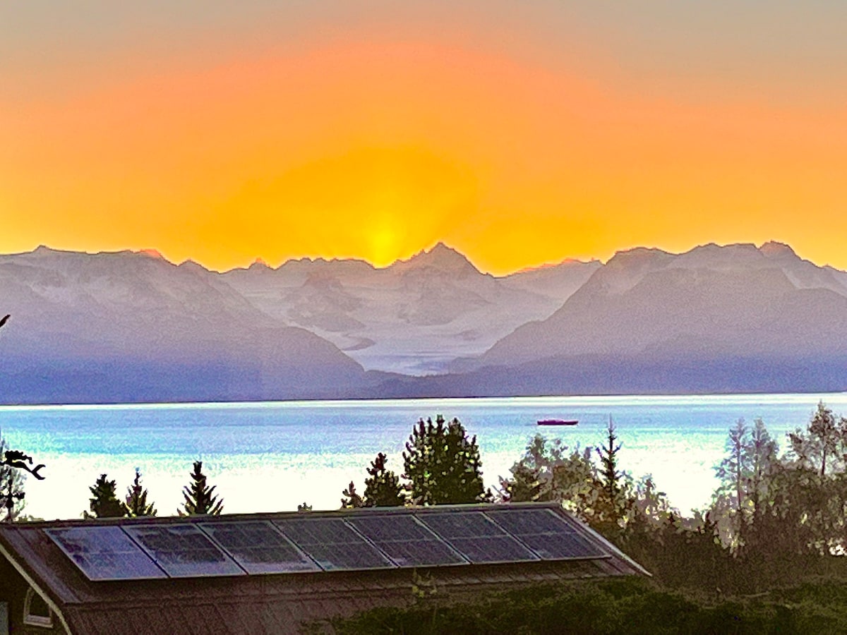 A panoramic view showcases a vibrant sunset over snow-capped mountains, with a serene bay reflecting orange and yellow hues. A large solar panel array is positioned in the foreground, complementing the natural landscape of trees and open water.