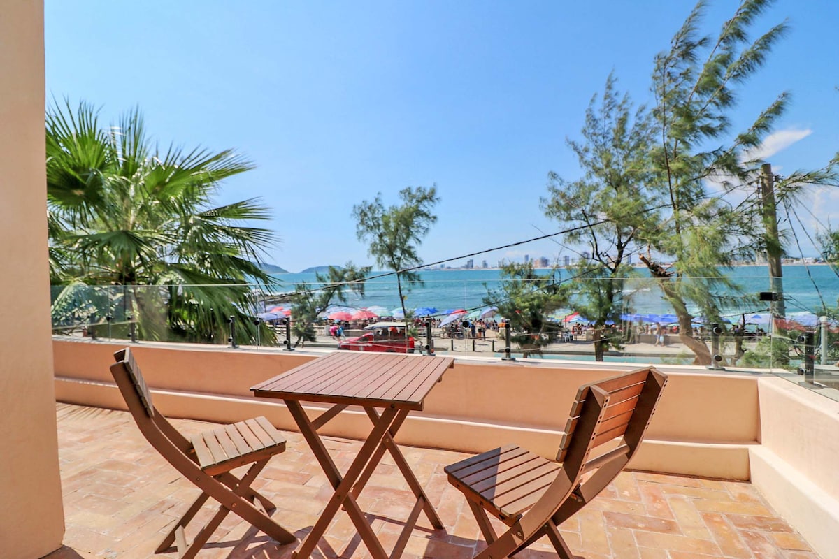 A sunlit terrace features a small wooden table surrounded by two matching chairs, offering views of the beach and ocean. The scene includes palm trees swaying gently in the breeze, while colorful umbrellas dot the sandy beach in the background.