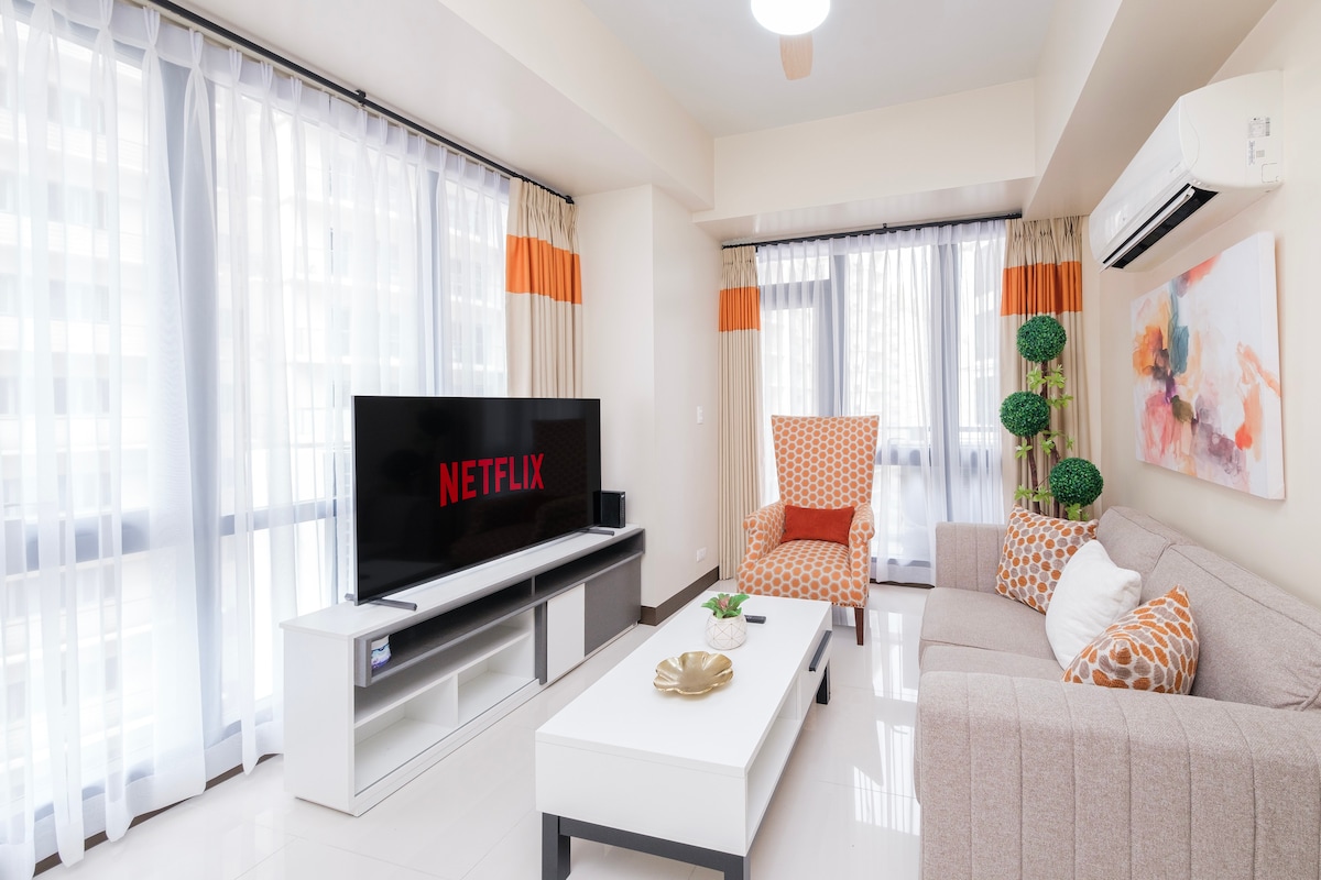 A bright living area features large windows draped with light sheer curtains, allowing ample natural light. A large television is mounted on a white media console, displaying the Netflix logo. Comfortable seating, including an accent chair with a geometric pattern, is arranged around a coffee table.