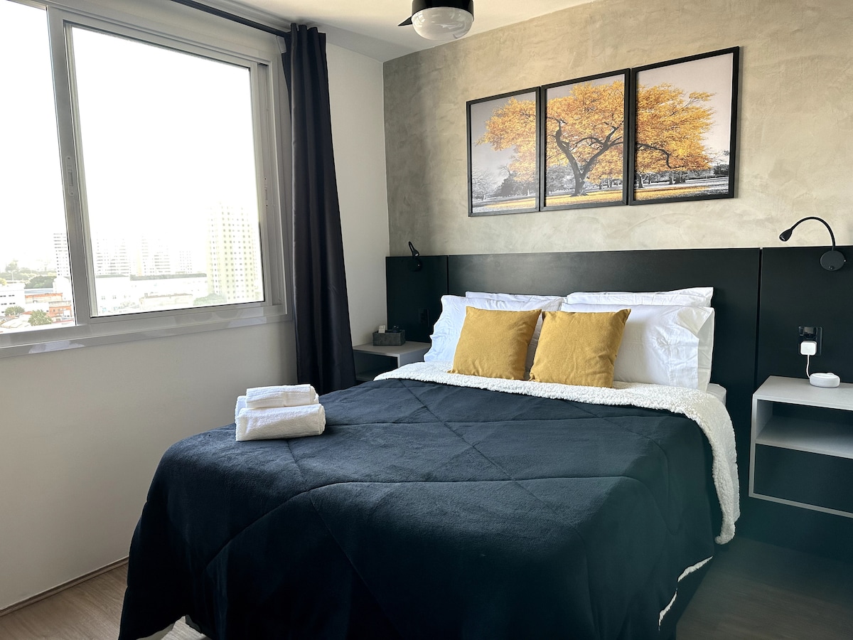 A spacious bed is layered with a dark blanket and accented by yellow pillows. Two framed photographs of trees are mounted above the bed. A bedside table is visible, along with a reading lamp. Large windows allow natural light to fill the room.