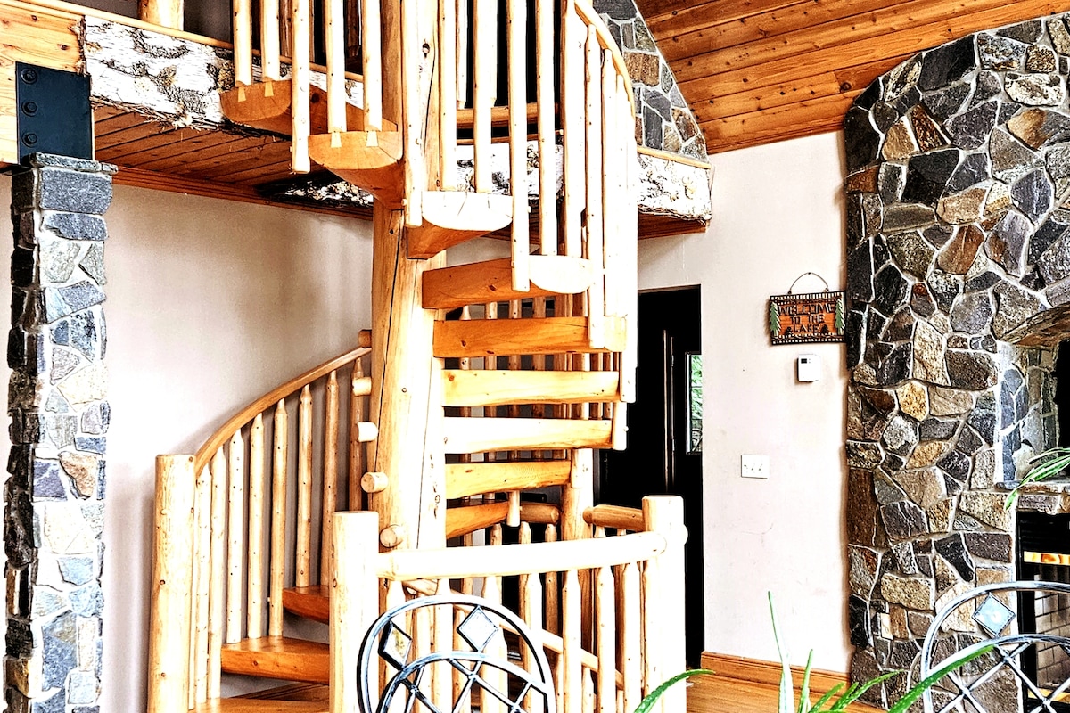 A wooden spiral staircase is featured prominently in a spacious room, surrounded by stone accents. The ceiling showcases natural wood paneling, and a light flows in from nearby windows. Black metal chairs are placed around a small dining table, enhancing the rustic charm of the space.