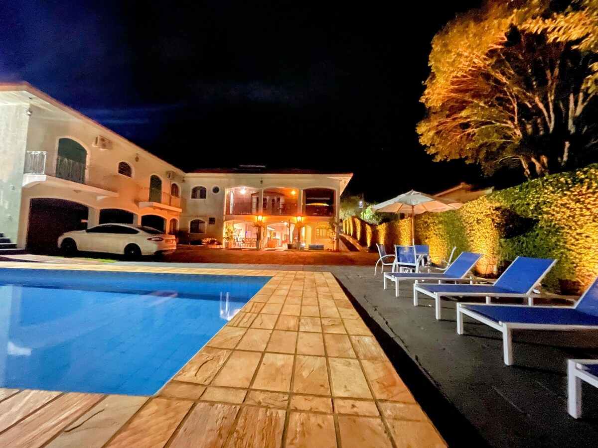 The image captures a tranquil outdoor pool area illuminated at night. White lounge chairs are positioned around the pool, and the surrounding greenery is softly lit. The two-story building in the background features large windows, revealing warm light from within.
