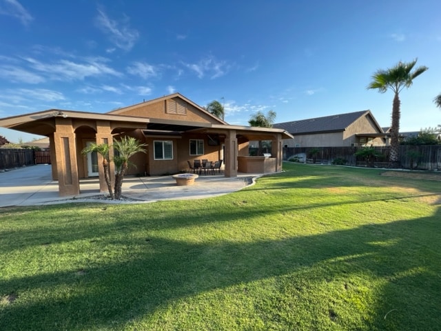 A spacious backyard is visible, featuring a covered patio with several seating options. The well-maintained grass area offers ample room for outdoor activities. Palm trees create a relaxed environment, while the home’s exterior includes large windows and an inviting roofline.