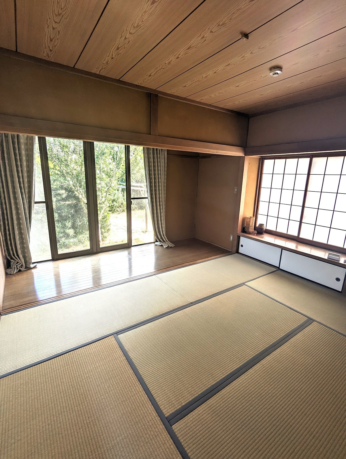 A traditional Japanese tatami room features large windows allowing natural light to fill the space. Smooth wooden floors complement the clean, minimalist decor. Floor cushions are present, while a shoji screen neatly frames the scenic view outside, enhancing the connection with nature.