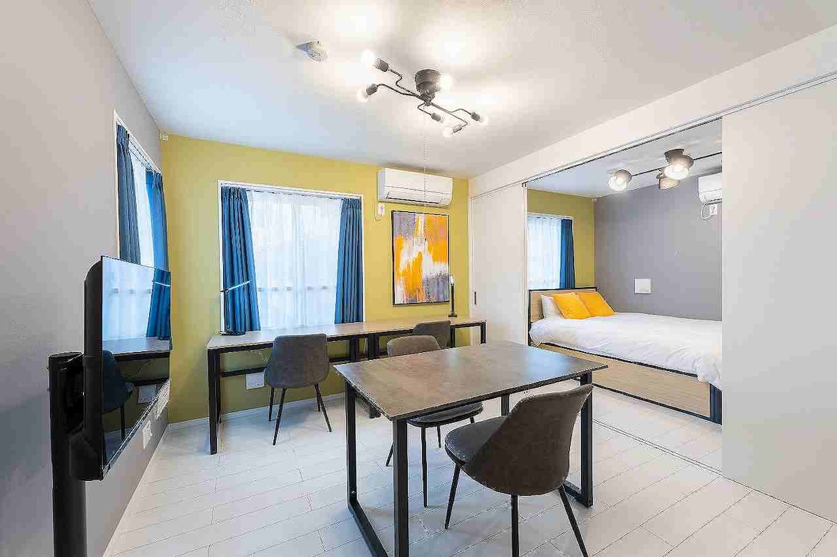 A modern living space is presented, featuring a dining table with three chairs and two windows with blue curtains allowing natural light. A comfortable bed, dressed in yellow and white linens, is visible in the background, while air conditioning units are mounted on the walls.