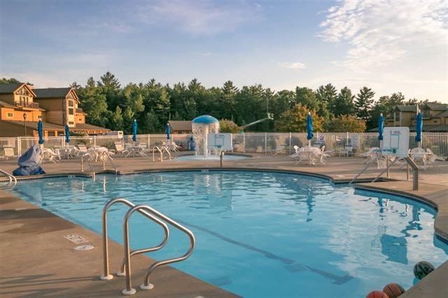 A swimming pool is surrounded by sun loungers and umbrellas, offering a refreshing retreat. A waterslide is visible at one end, and a wooden fence provides a sense of privacy. Lush greenery lines the background, enhancing the serene atmosphere.