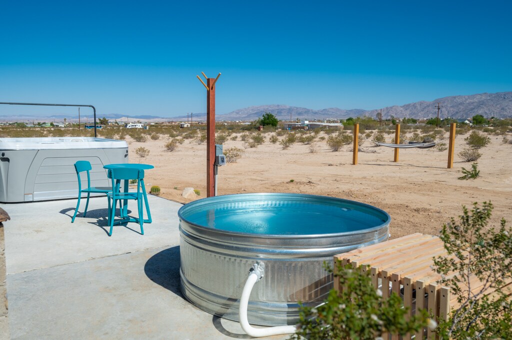 A cowboy pool sits adjacent to a hot tub on a concrete patio, surrounded by the desert landscape. Two blue chairs are positioned nearby, offering a place to relax and enjoy the view of the expansive, sunlit terrain.